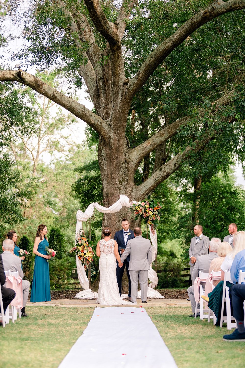 Outdoor wedding ceremony under a large tree with white aisle runner and floral arch.