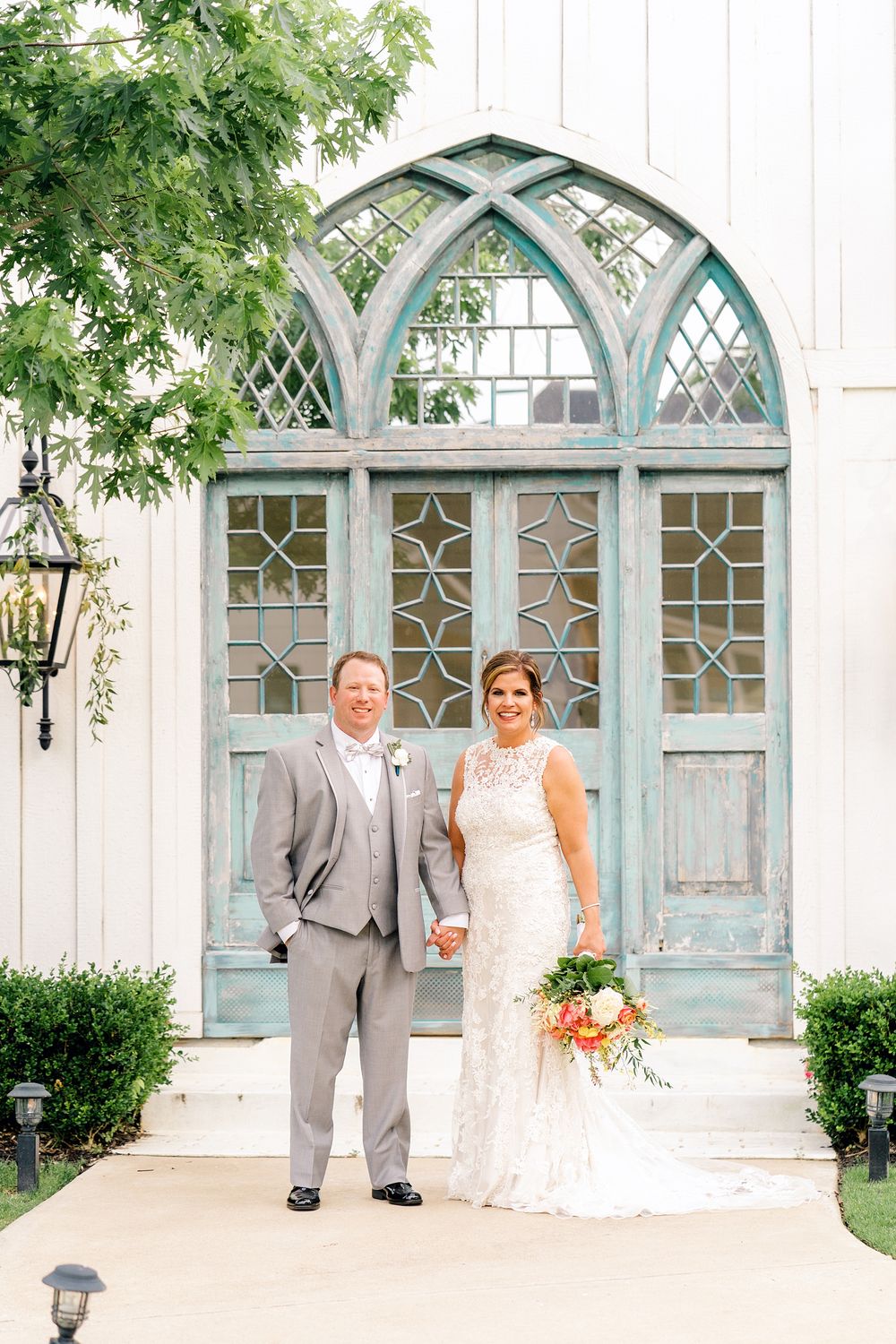A wedding couple poses in front of a beautiful turquoise arched church door surrounded by greenery.