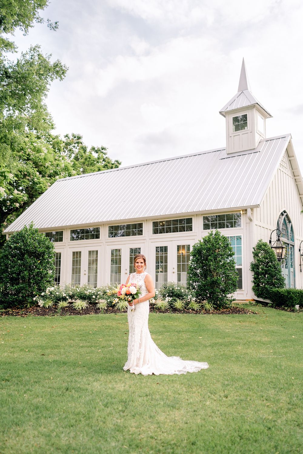 A white chapel with spire stands behind a manicured lawn and garden on an overcast day.