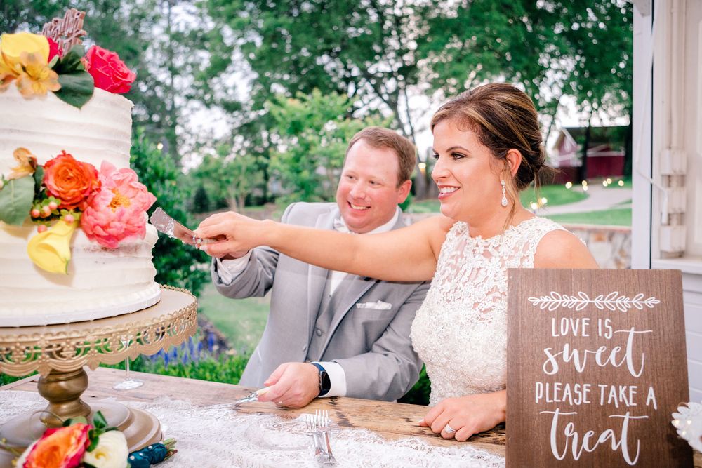 A couple cuts their elegant tiered wedding cake outdoors with a rustic wooden sign displayed nearby.