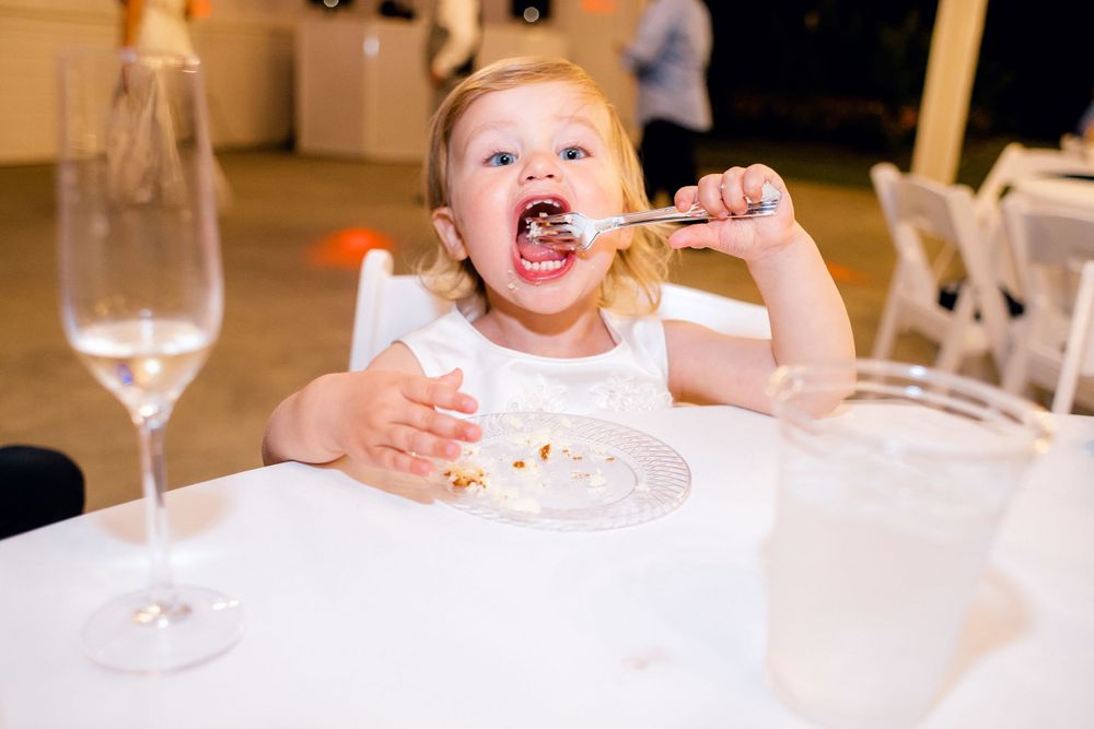 An excited small child eating cake at a formal event with a wine glass on the table.