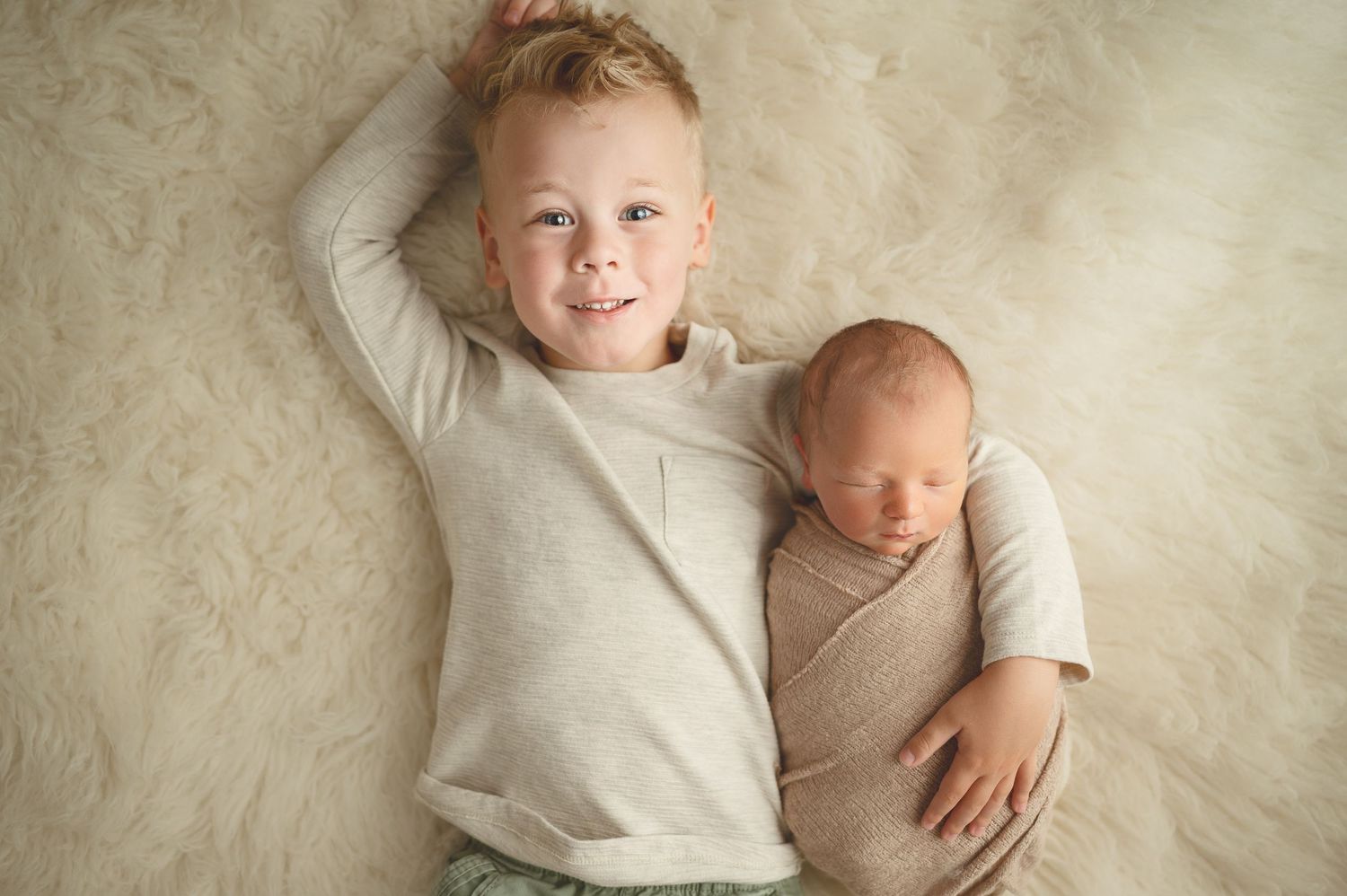 Two children in light-colored clothing lying on a cream-colored backdrop during a photo session.