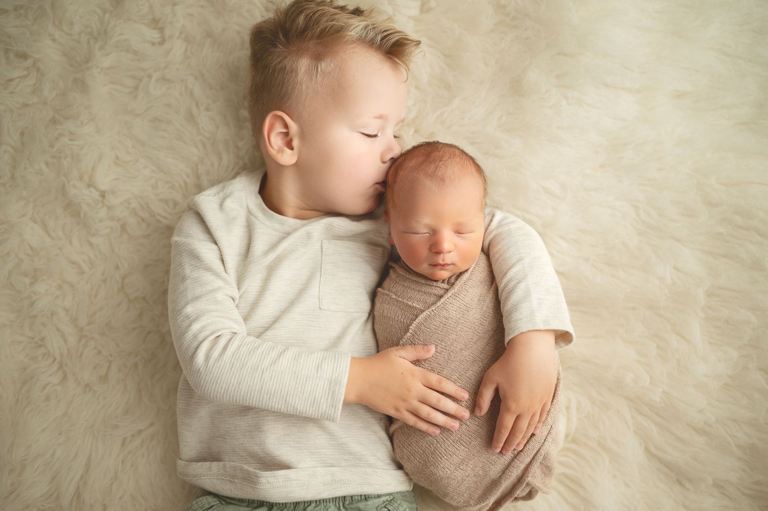 Siblings sharing a tender moment while lying on a cream-colored background.
