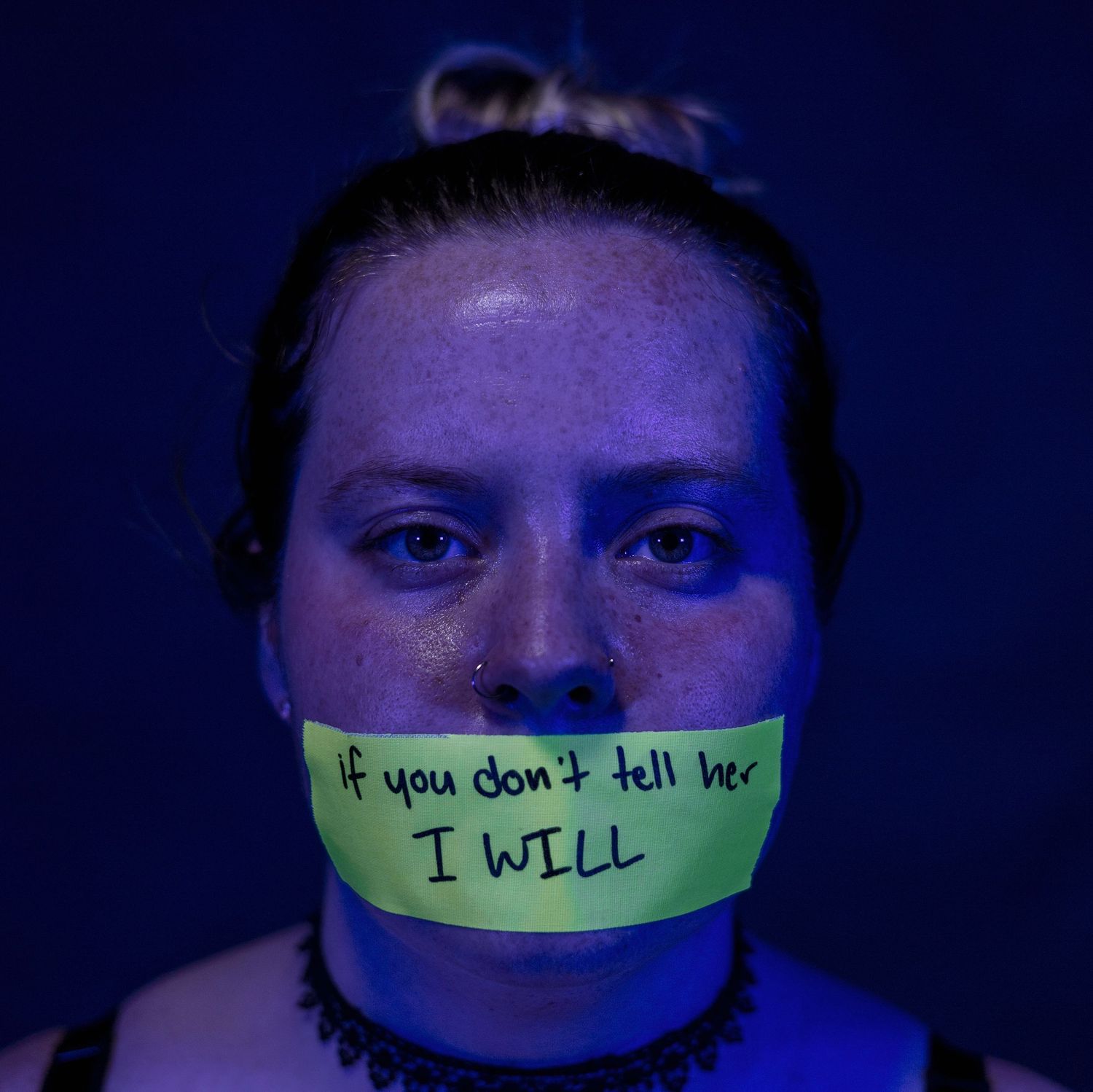 Person with neon-lit face wearing tape over mouth with text message written on it.