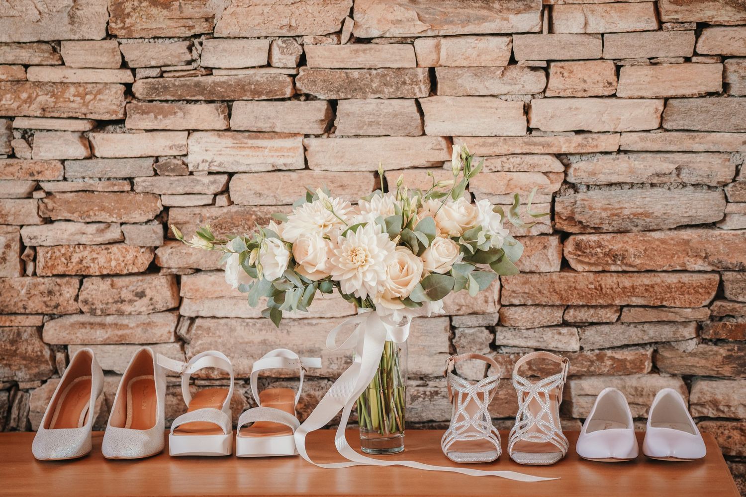 Collection of bridal shoes and floral arrangement displayed against brick wall.