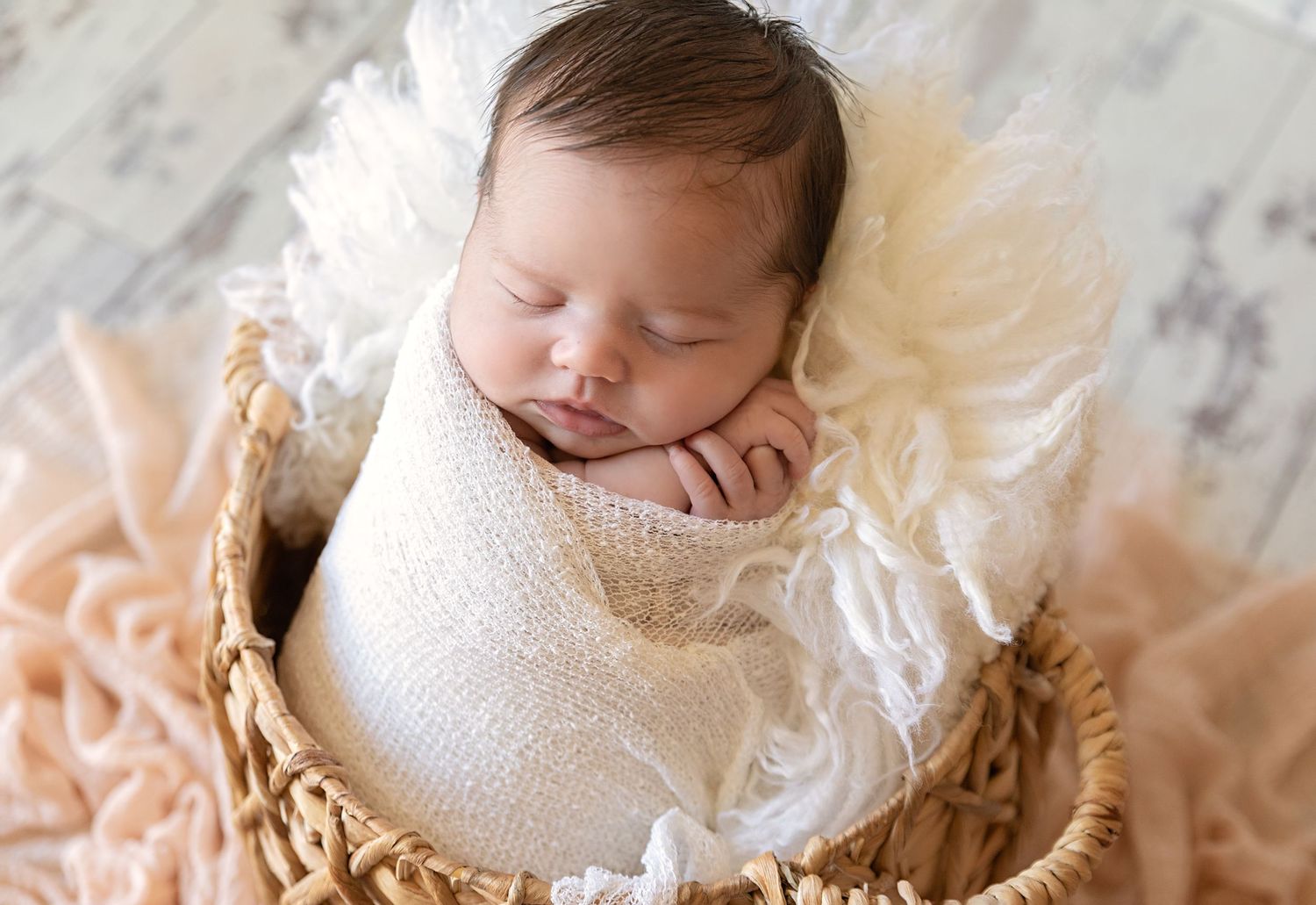Peaceful newborn sleeping in cream wrap inside woven basket on textured background.