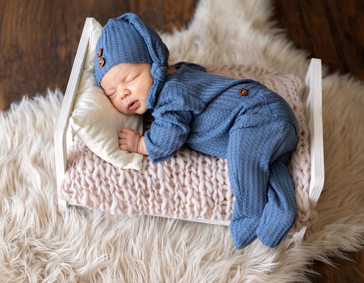Sleeping baby in blue knit outfit resting on miniature bed with plush bedding.