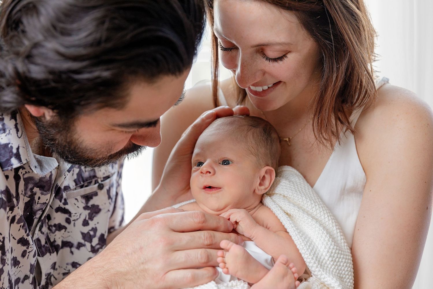 A tender family moment as parents share a loving gaze with their newborn baby wrapped in white.
