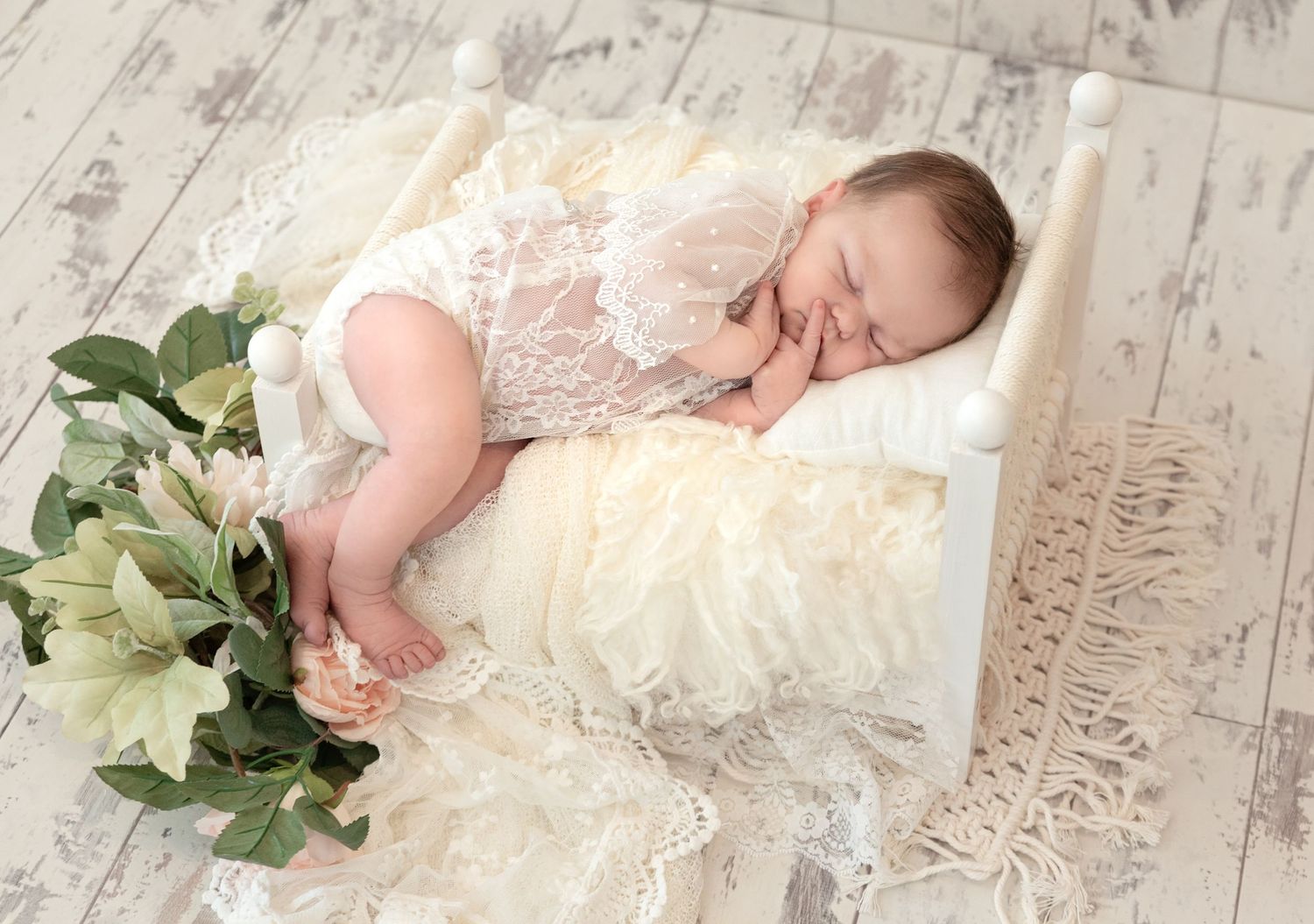 Sleeping newborn in lace outfit on vintage wood floor with white flowers.