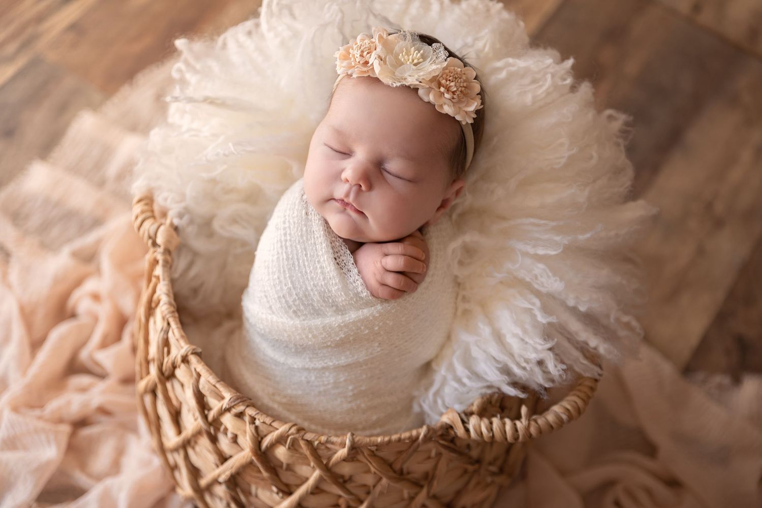 Sleeping newborn in white wrap and peach bow resting in woven basket.
