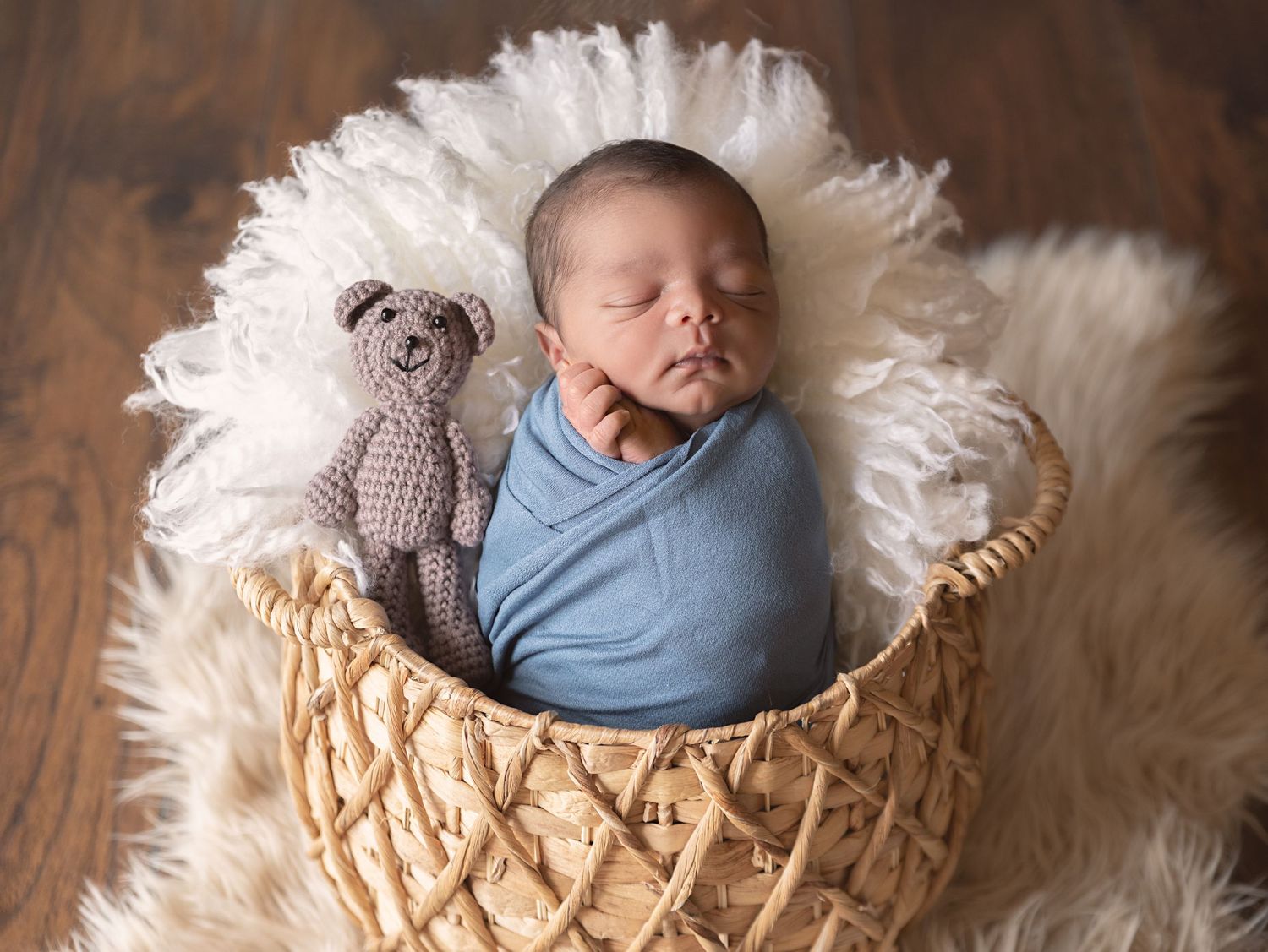 Sleeping newborn wrapped in blue swaddle with knit toy in woven basket on white fur.