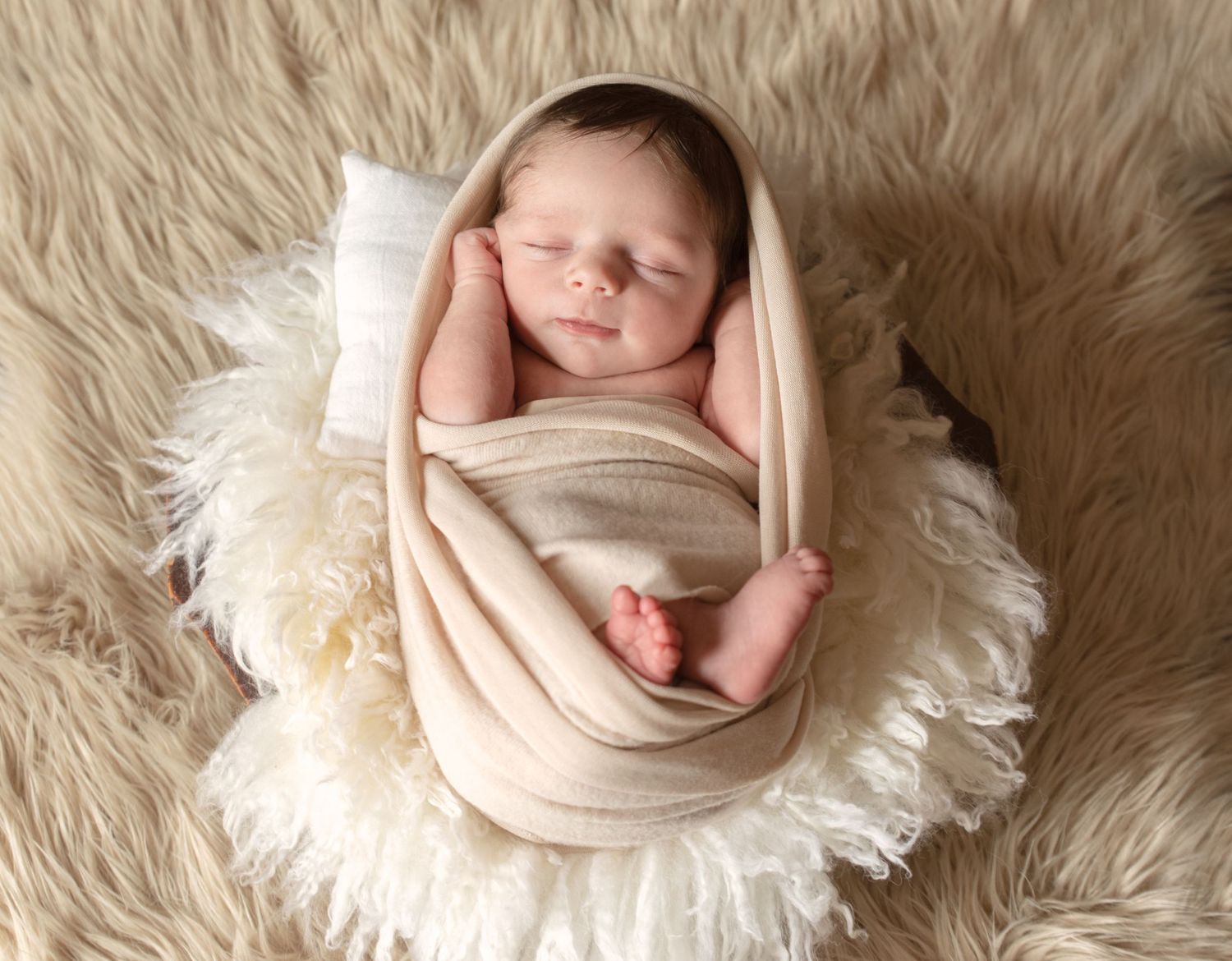 Peaceful newborn sleeping on cream fur rug wrapped in beige swaddle.
