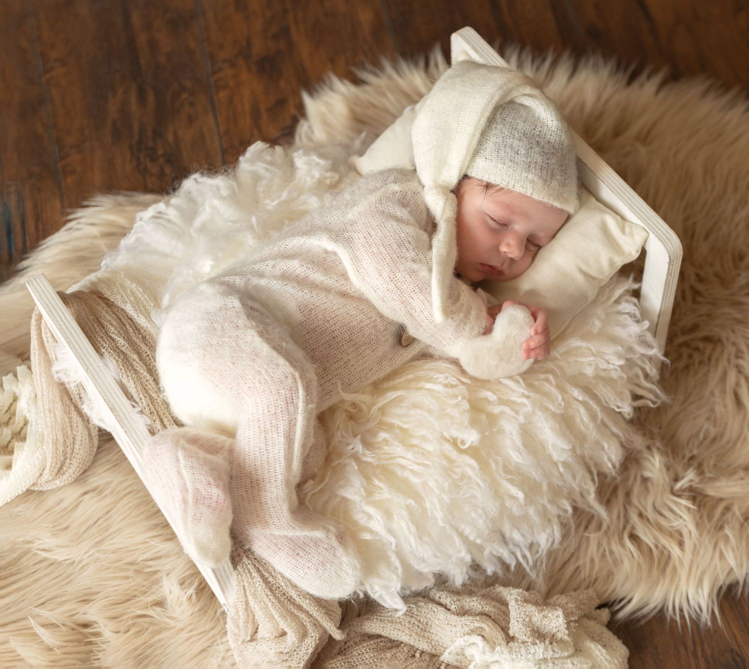 Sleeping baby wrapped in cream knit blanket lying on fluffy white fur rug.