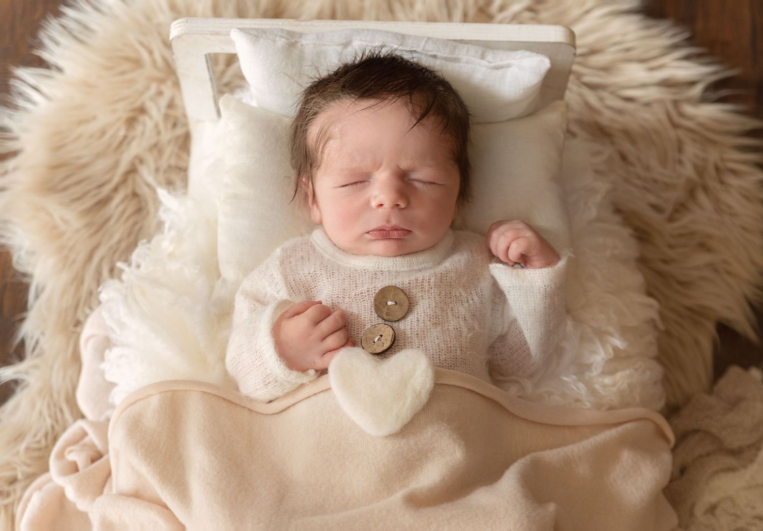 Newborn baby sleeping peacefully on beige fur blanket wearing white knit outfit.