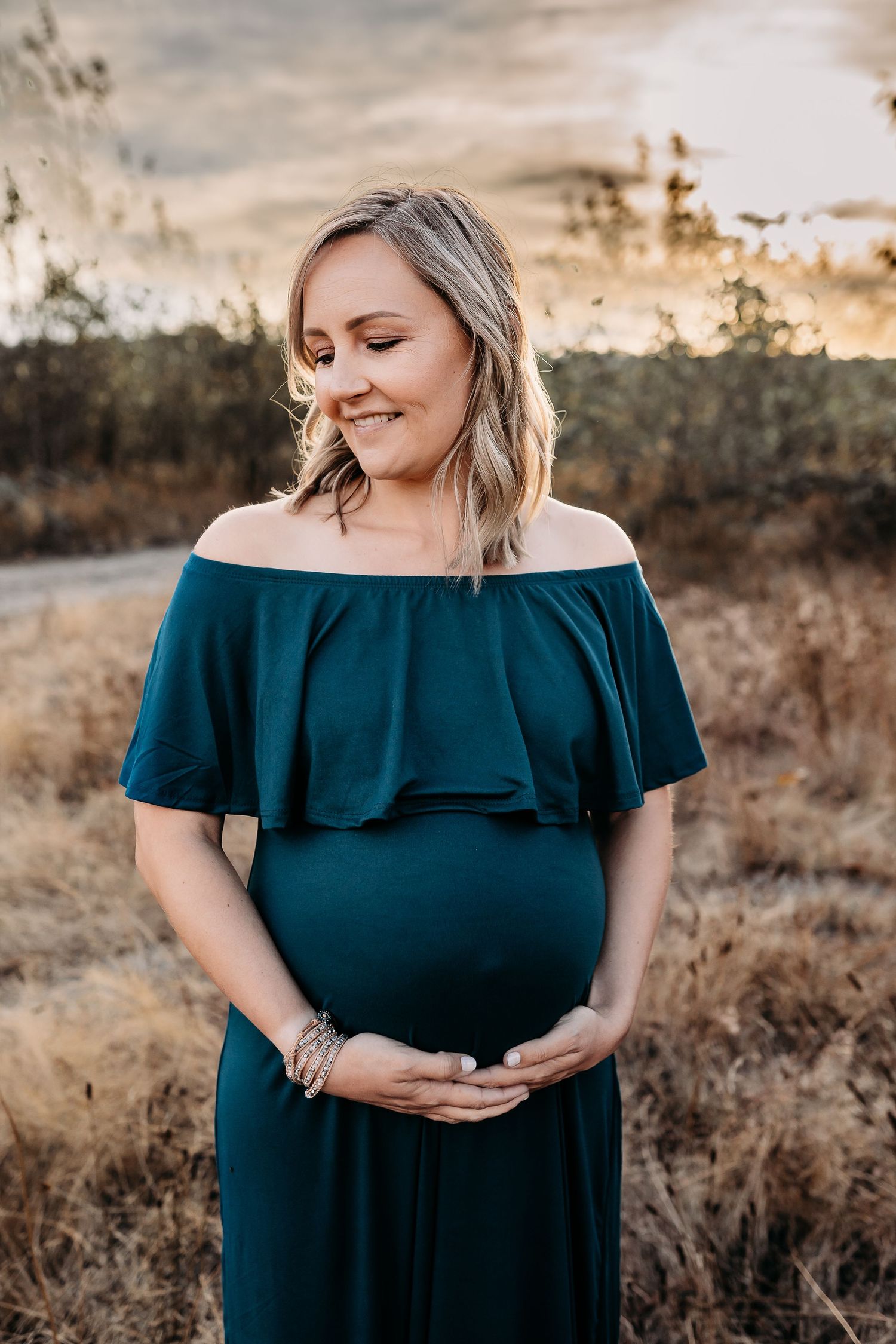 Tami smiles with her hands cradling her pregnant belly in maternity dress with a warm, summer sunset