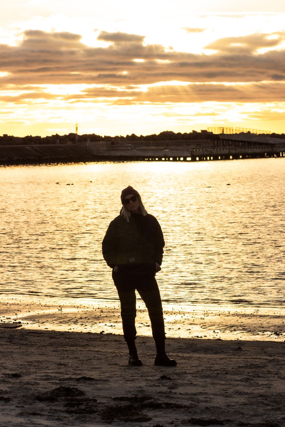 Lone figure in silhouette against golden sunset waters and distant pier.