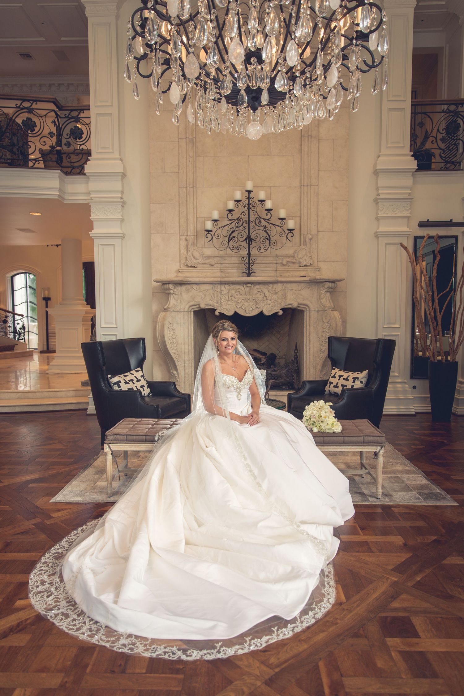 A bride in a white gown sits elegantly under a crystal chandelier in a luxurious ballroom setting.