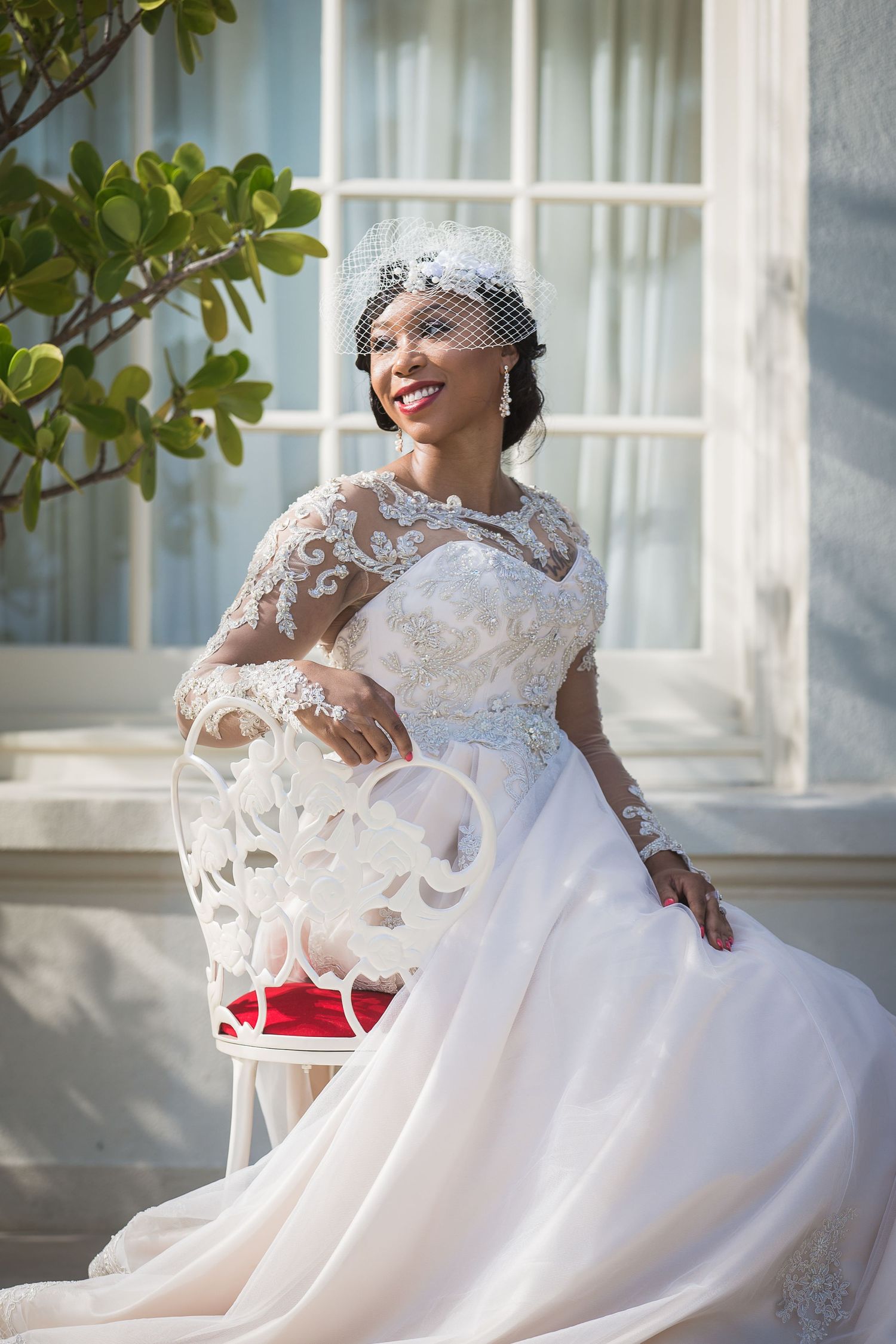 Sunlit bridal portrait featuring a flowing white wedding gown against white windows and green foliage.