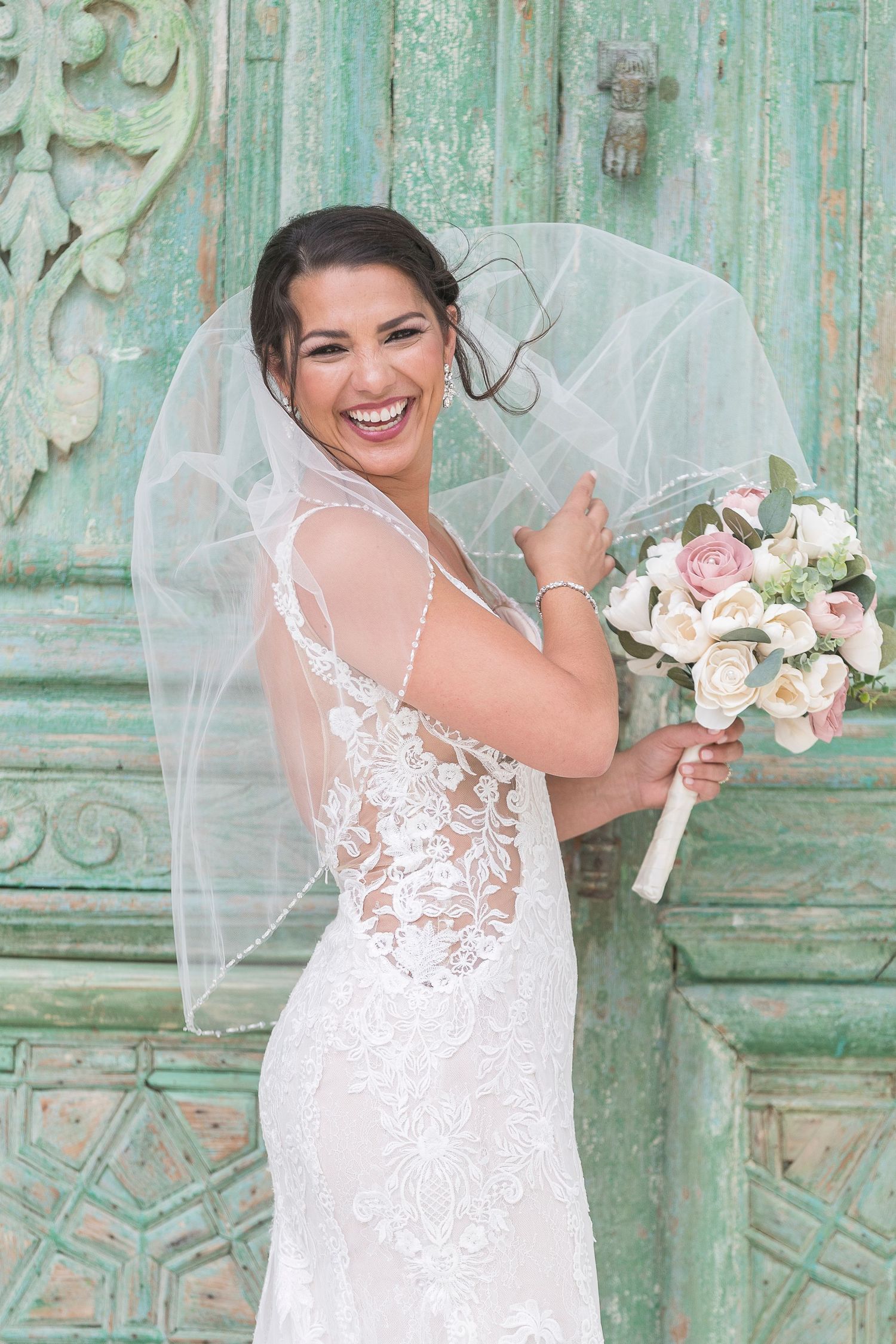 A bride in a lace wedding dress poses against a vintage mint green door while holding a pink and white bouquet.