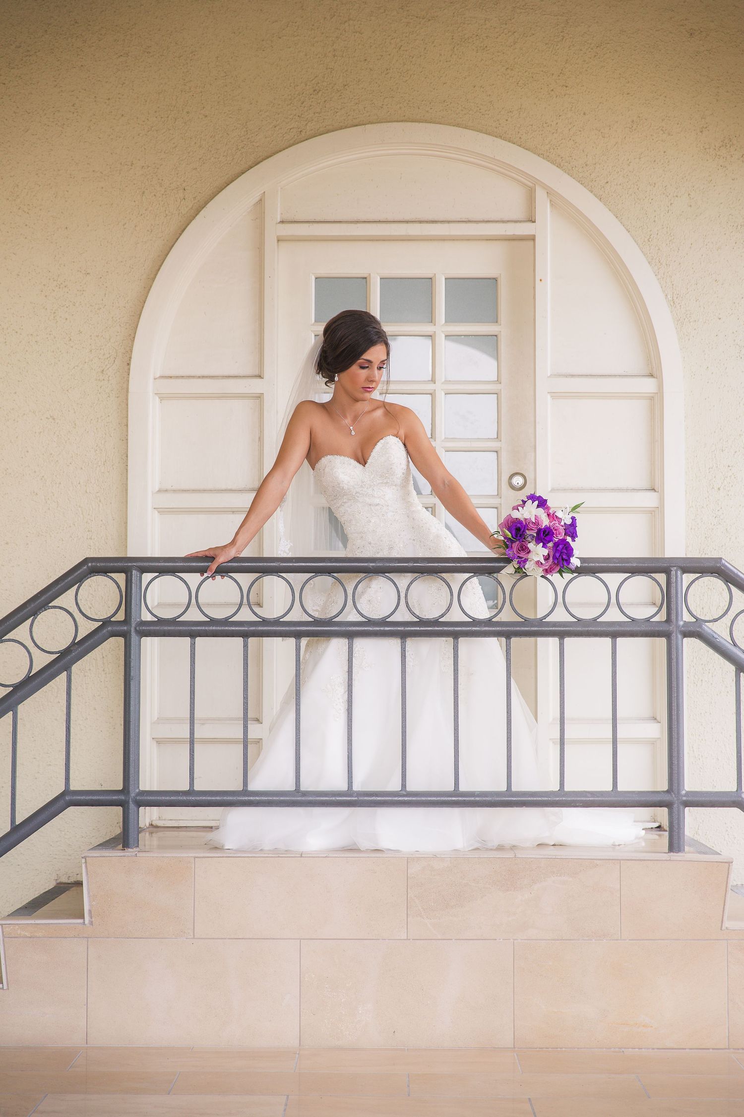 A bride poses along an elegant balcony railing with arched windows and purple florals.