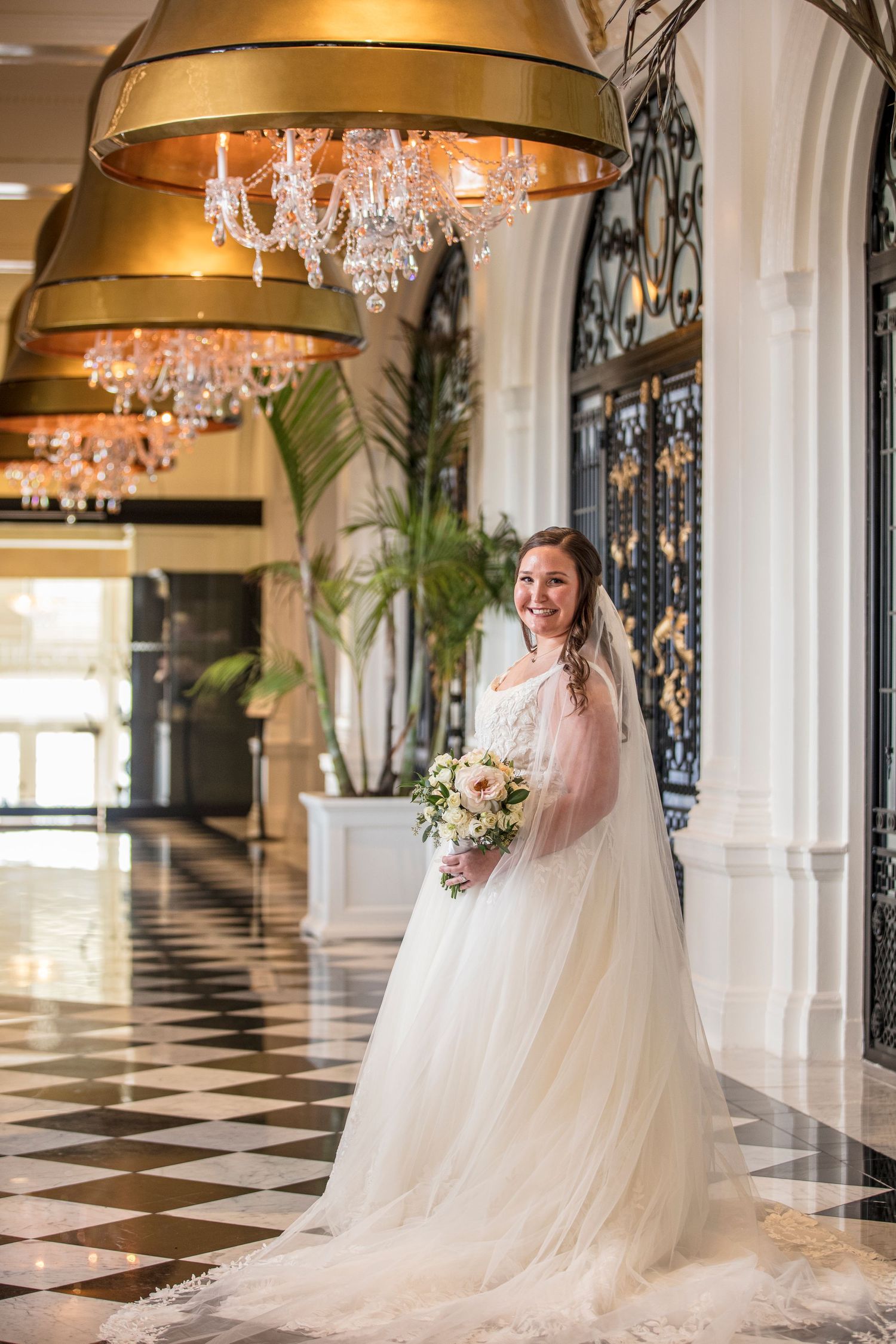 Elegant bridal portrait in grand hallway with crystal chandeliers and checkered floor.