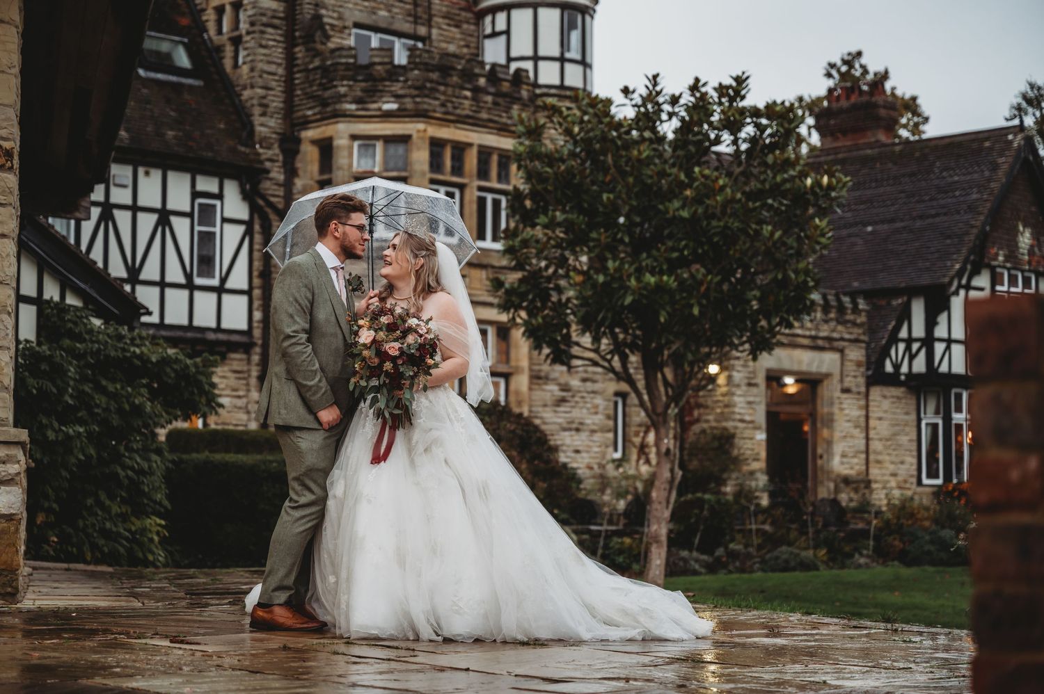 A romantic wedding portrait in front of a Tudor-style stone mansion with autumn foliage.