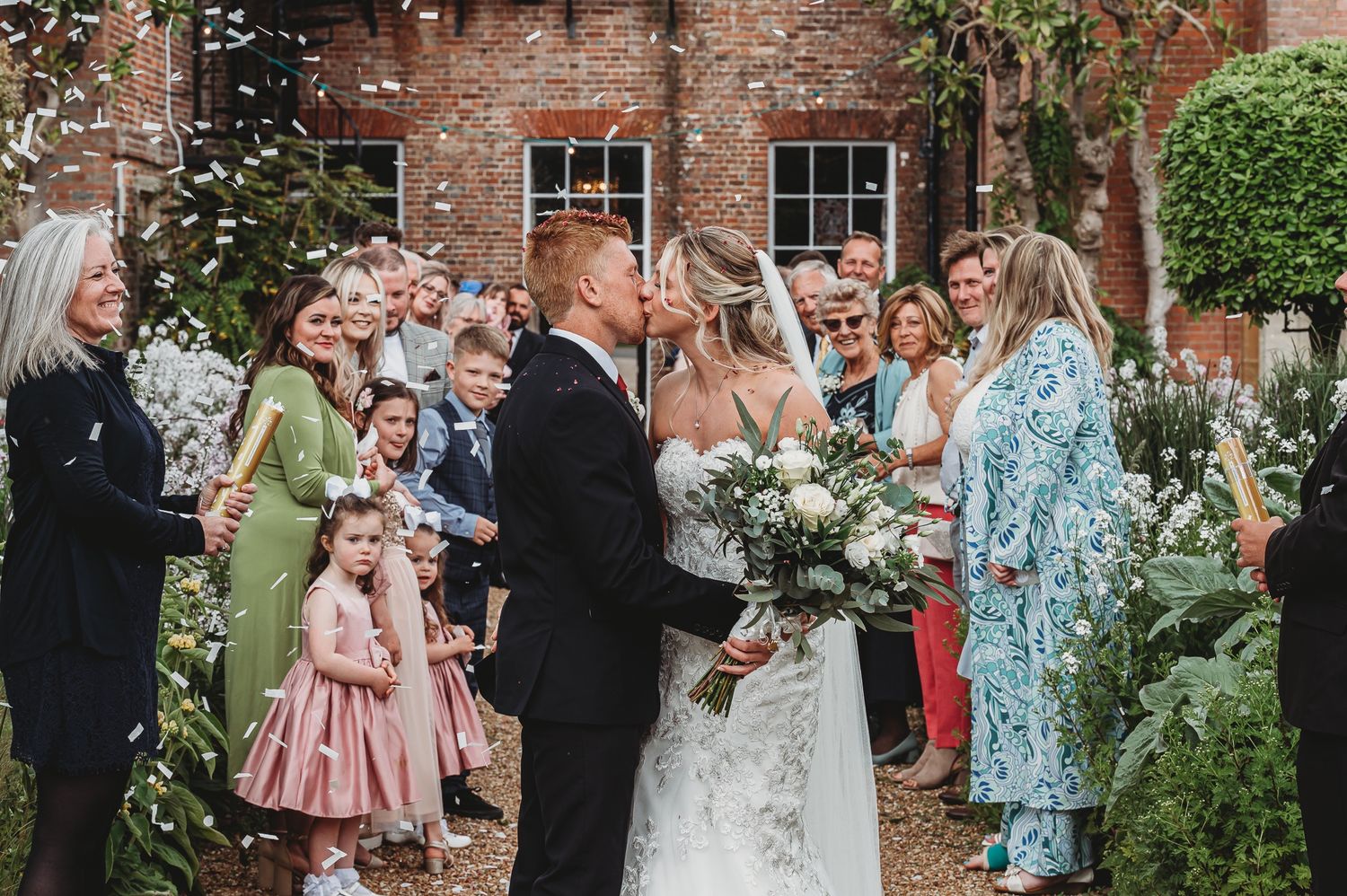 A joyful outdoor wedding ceremony with guests throwing confetti near a brick building.