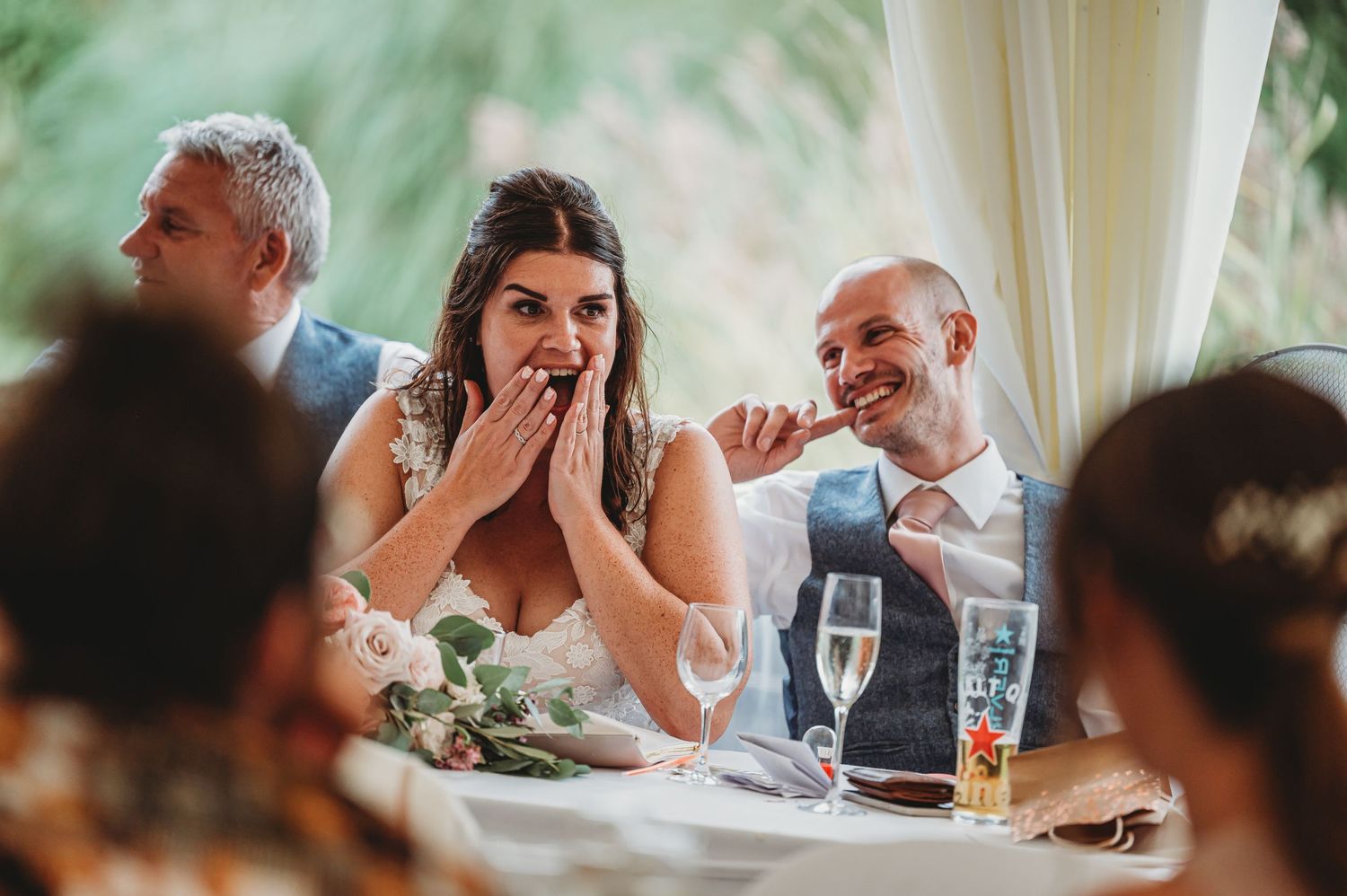 Wedding reception candid moment showing joyful reactions during speeches at a decorated dining table.