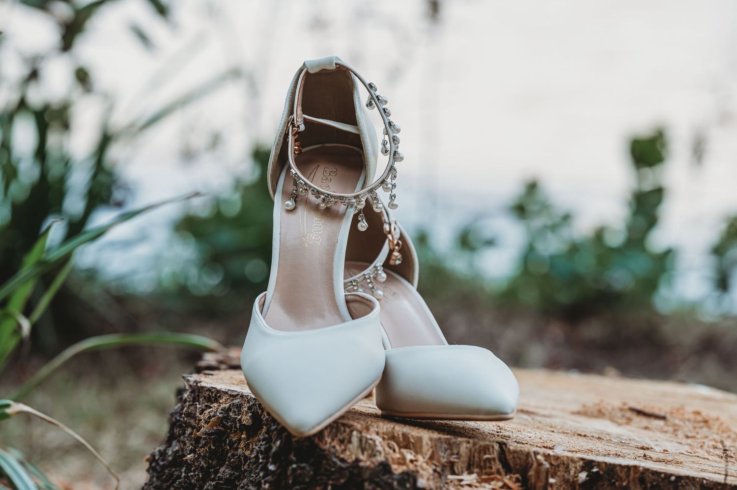 White bridal heels with delicate straps photographed on rustic wooden log outdoors.