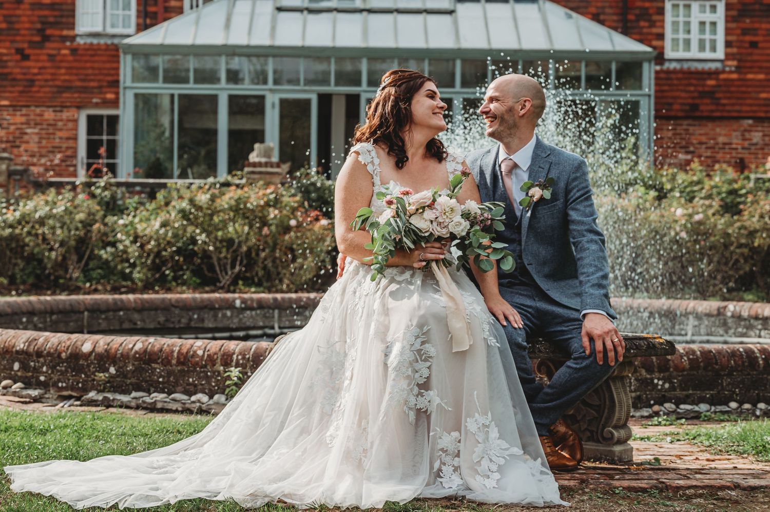 Newlyweds share a joyful moment in front of a red brick conservatory garden setting.