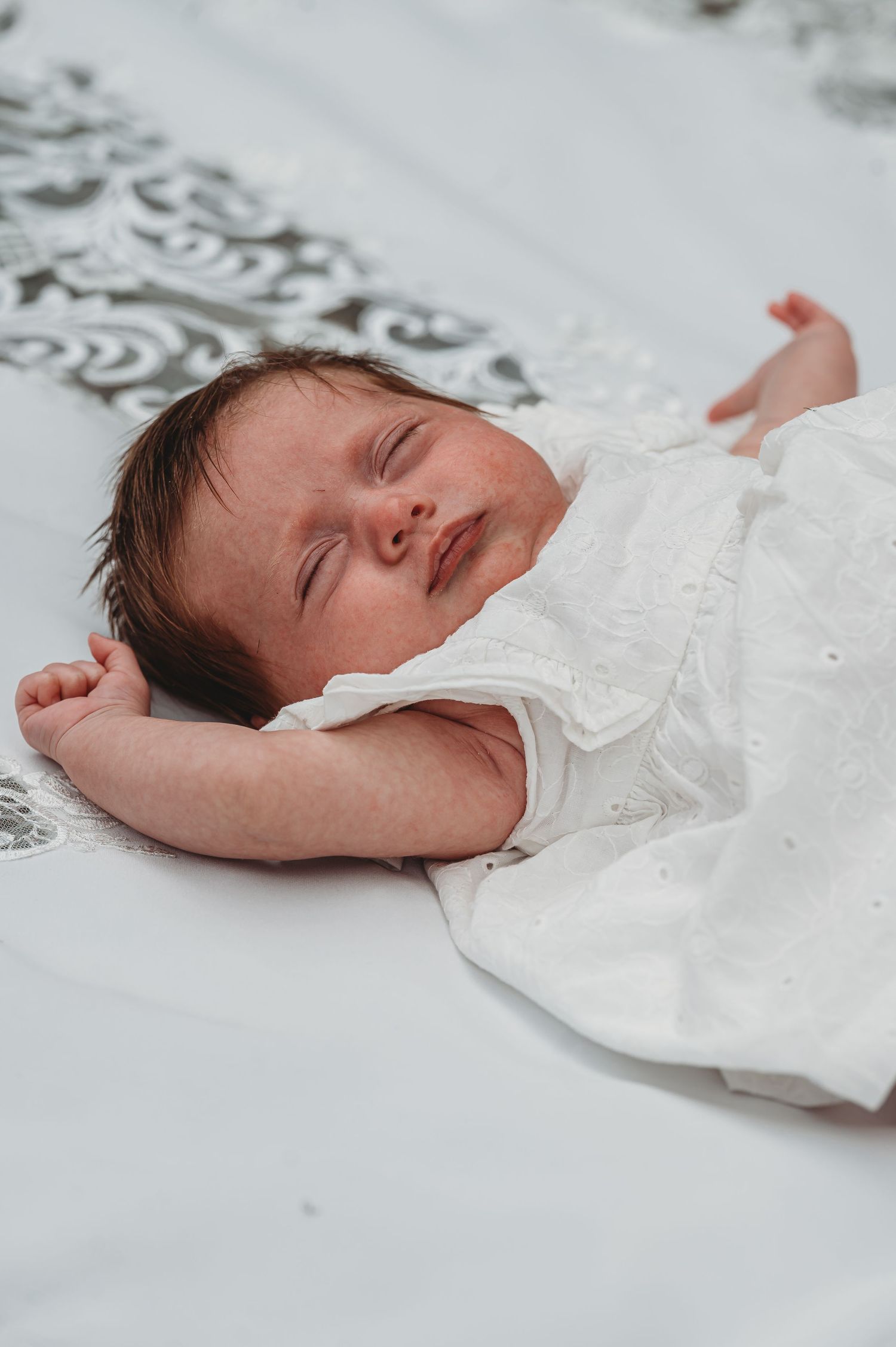 Peaceful sleeping newborn in white lace outfit resting on patterned bedding.