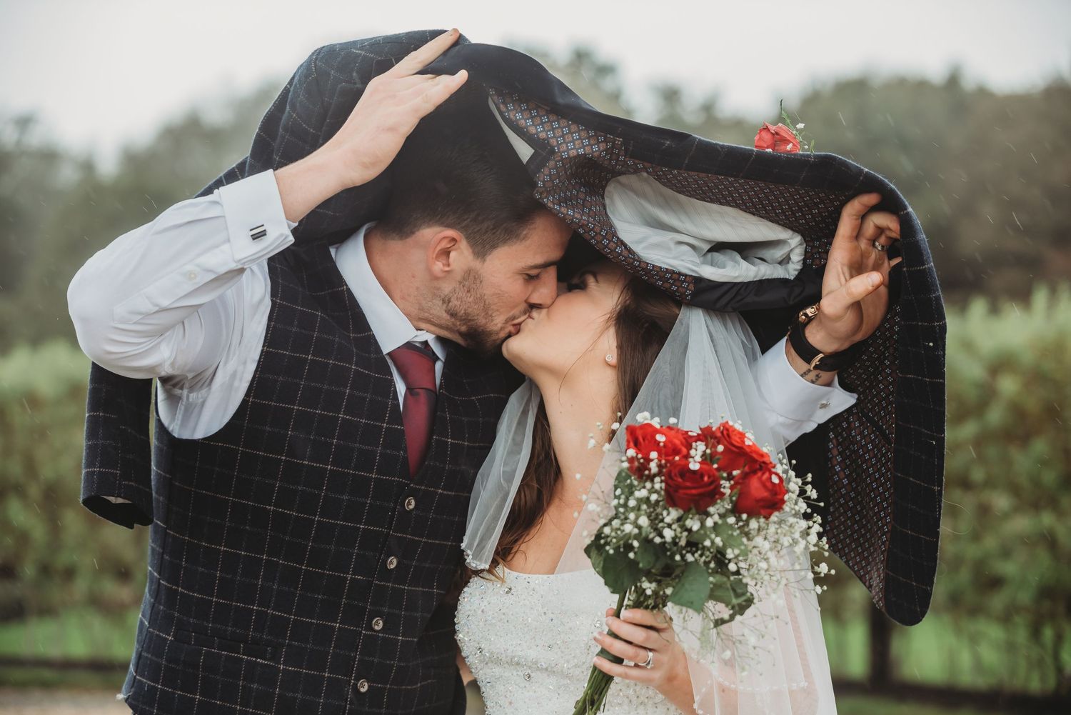 Romantic newlyweds share an intimate moment under a veil while holding red flowers in an outdoor garden setting.