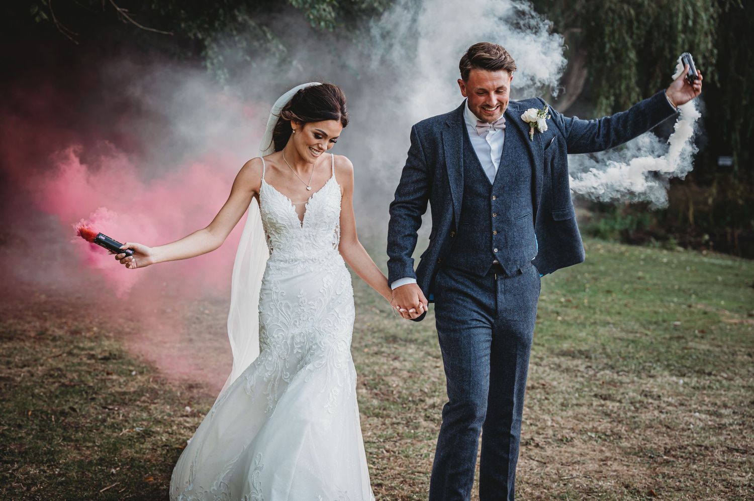 Wedding couple holds hands while walking with pink smoke bomb creating dramatic outdoor photo effect.