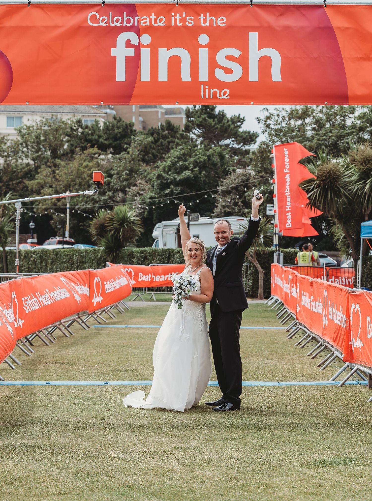 Wedding couple celebrates under a finish line banner at an outdoor event.