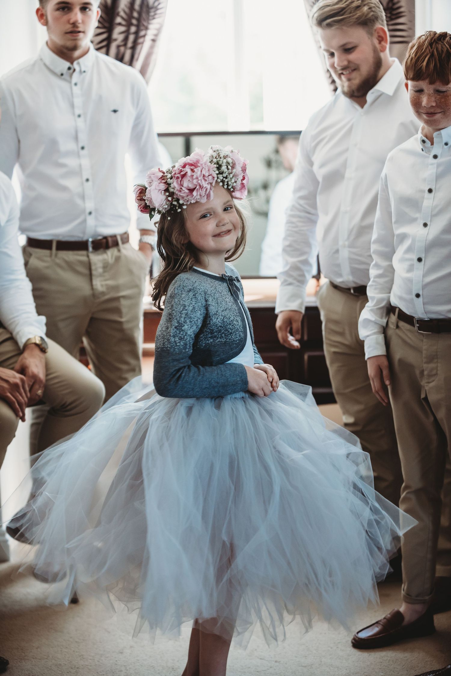 Young flower girl in blue tulle dress and floral crown surrounded by formal-dressed attendants.