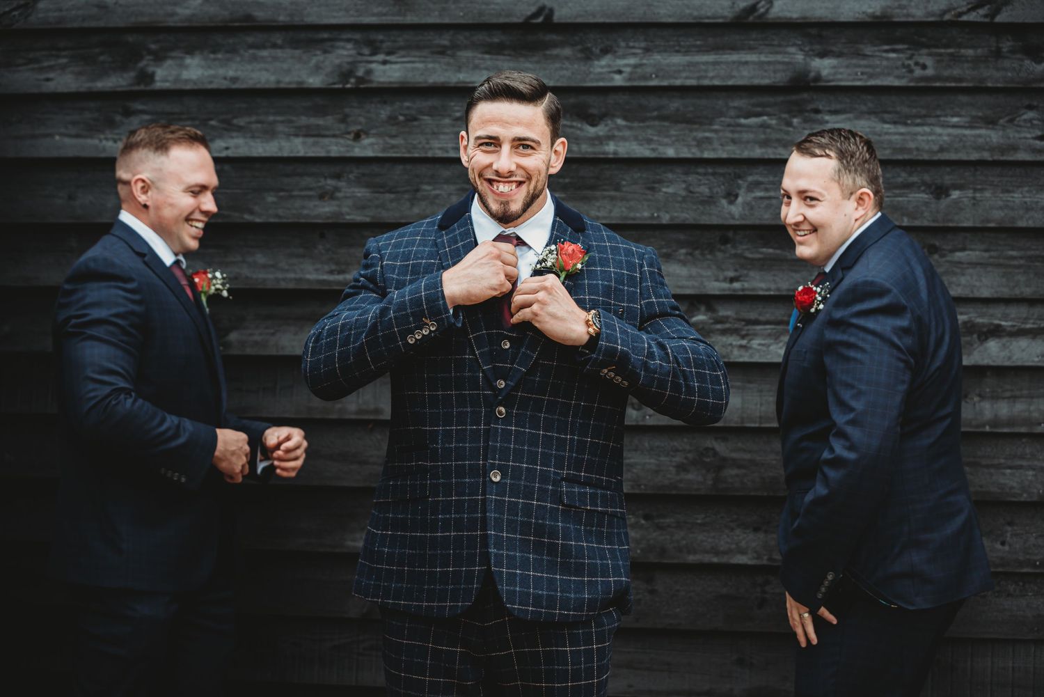 Wedding party celebrates with groomsmen in checkered suits against rustic wood wall.