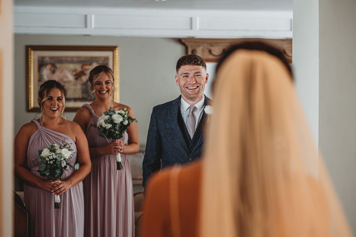 Wedding party members in mauve dresses react emotionally as they watch someone during a pre-ceremony moment.