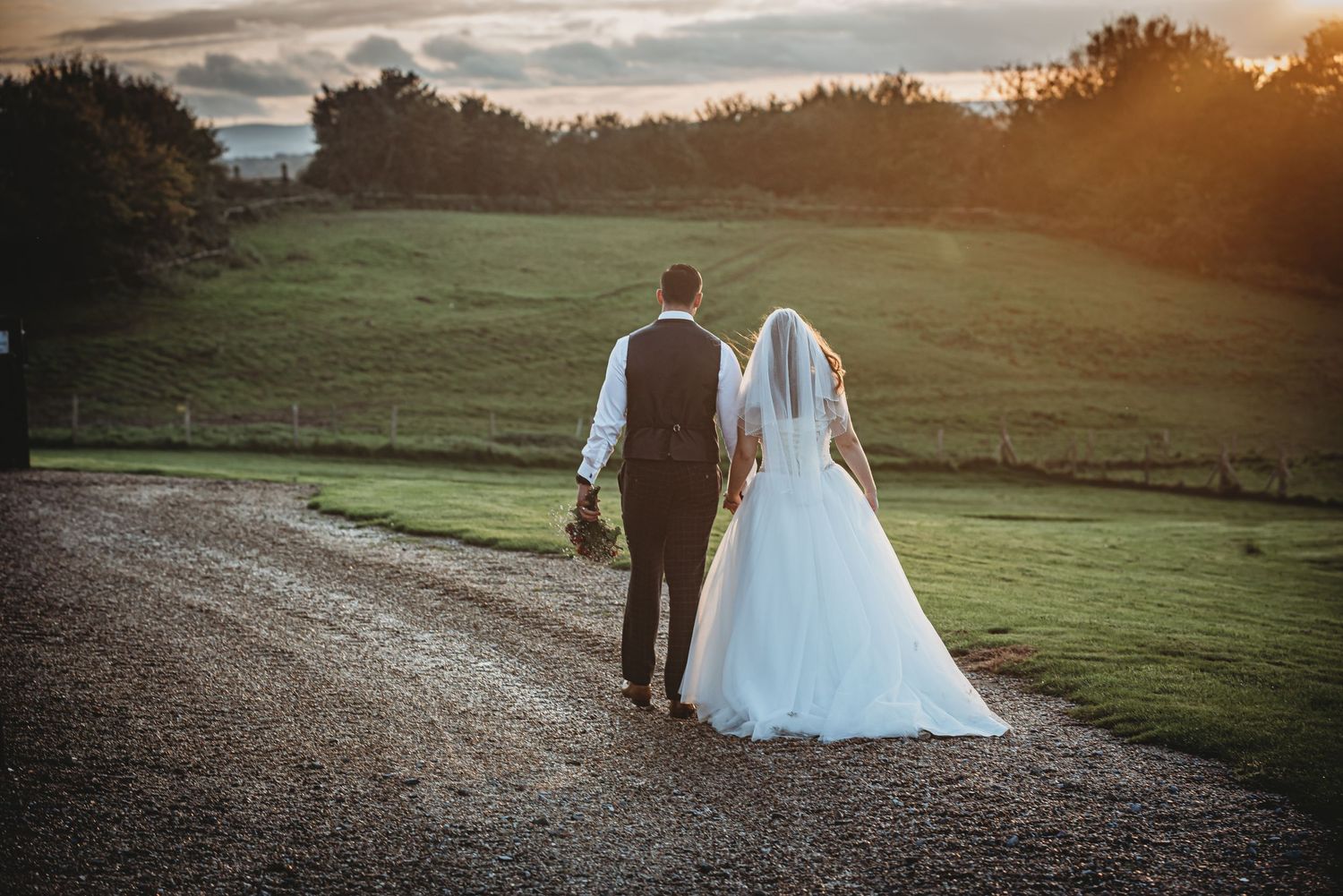 A newlywed couple walks down a gravel path at sunset with rolling green hills in the background.