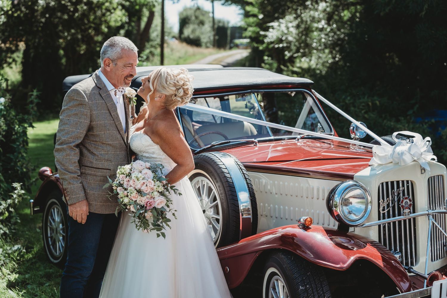 Newlyweds pose beside a vintage red car decorated with white ribbons.