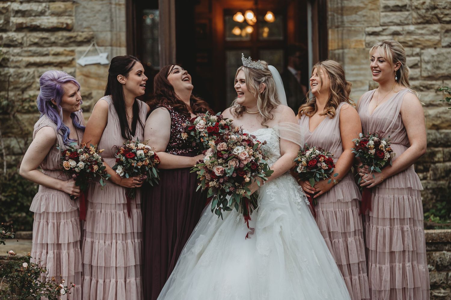 Bridesmaids in pink dresses share a joyful moment with the bride outside a stone building.