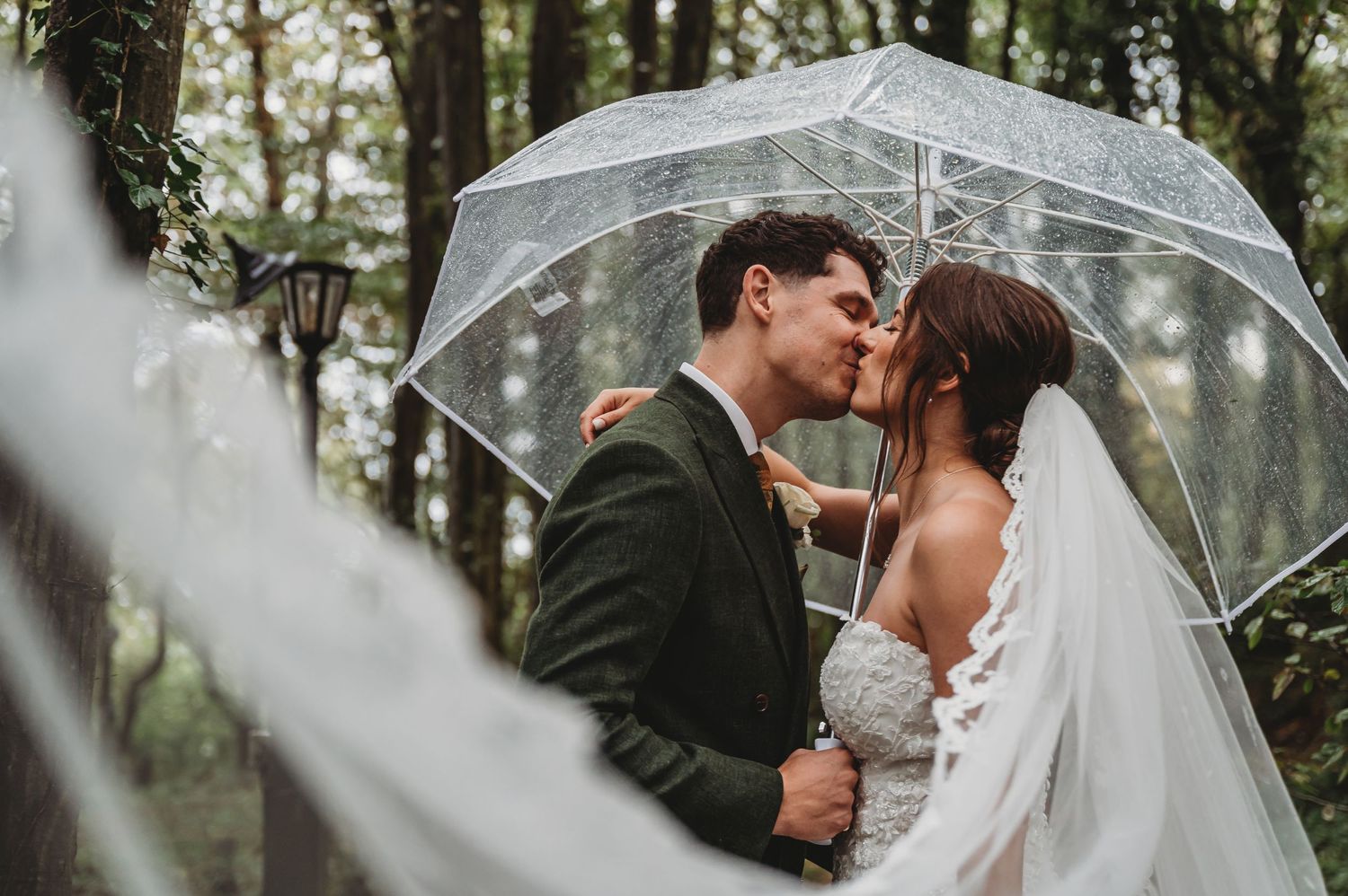 Romantic rainy day wedding portrait under a clear umbrella in a wooded setting.