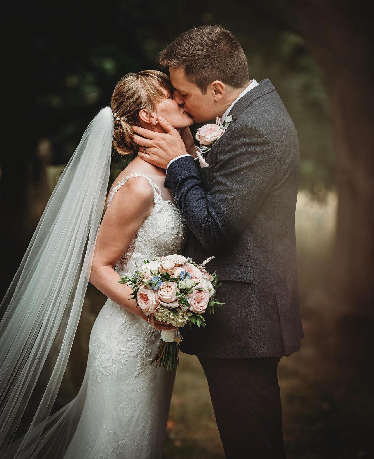 A romantic wedding couple shares an intimate kiss while holding a pink and white floral bouquet.