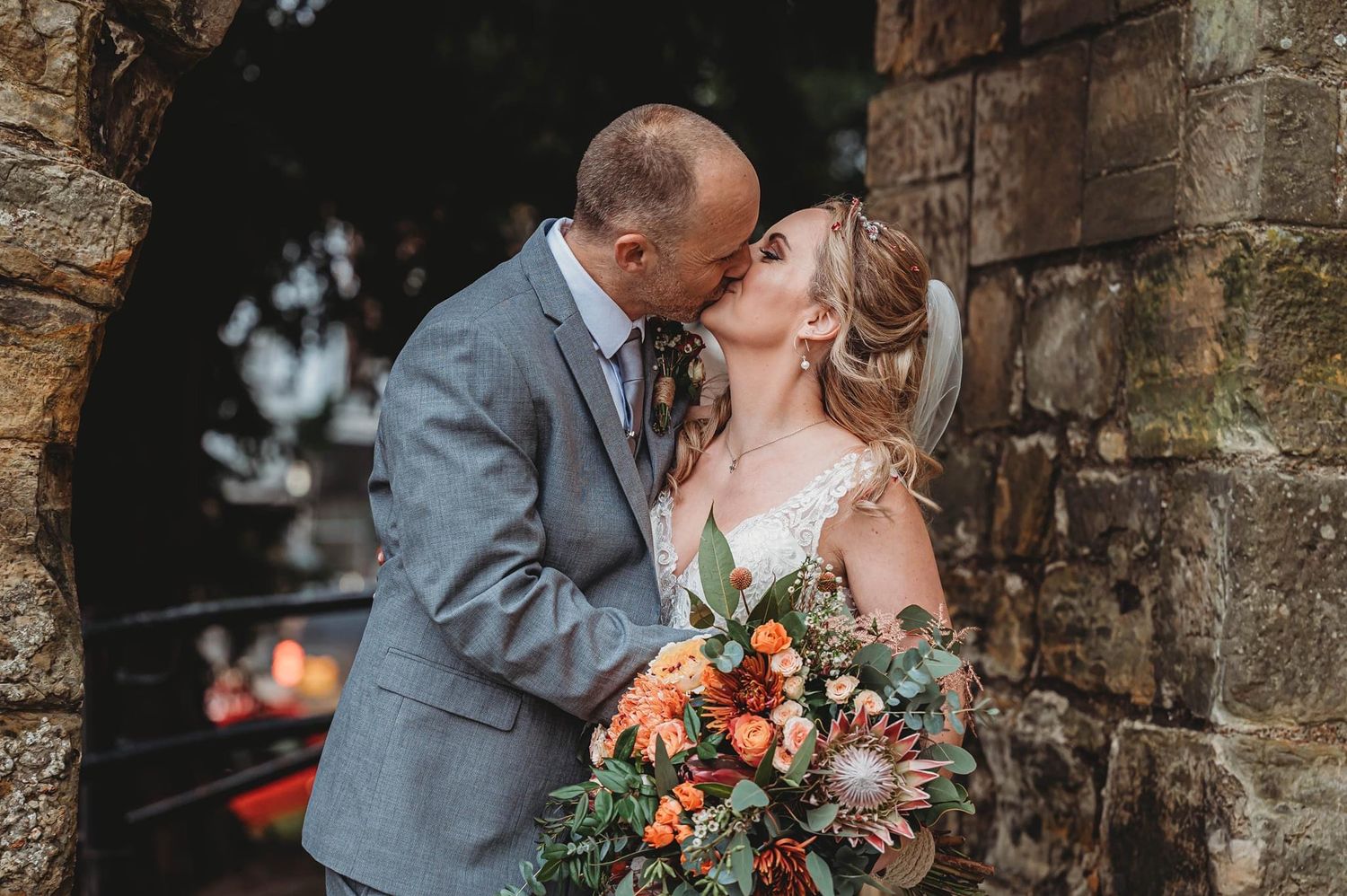Autumn wedding portrait against stone archway with vibrant orange and rust colored bouquet.