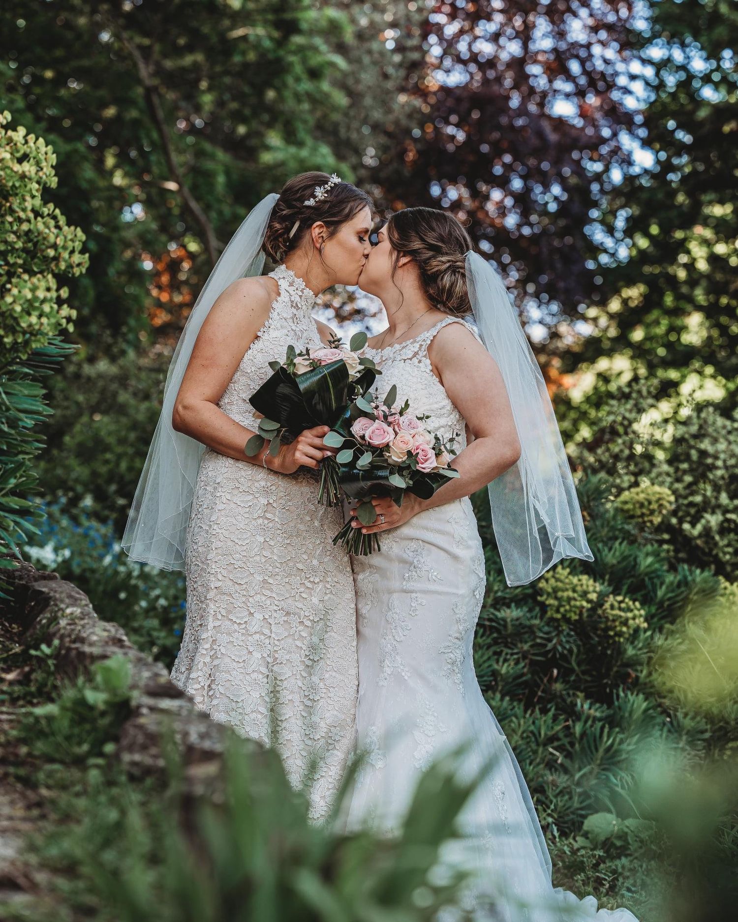 Two brides share an intimate moment in a lush garden setting while holding beautiful floral bouquets.