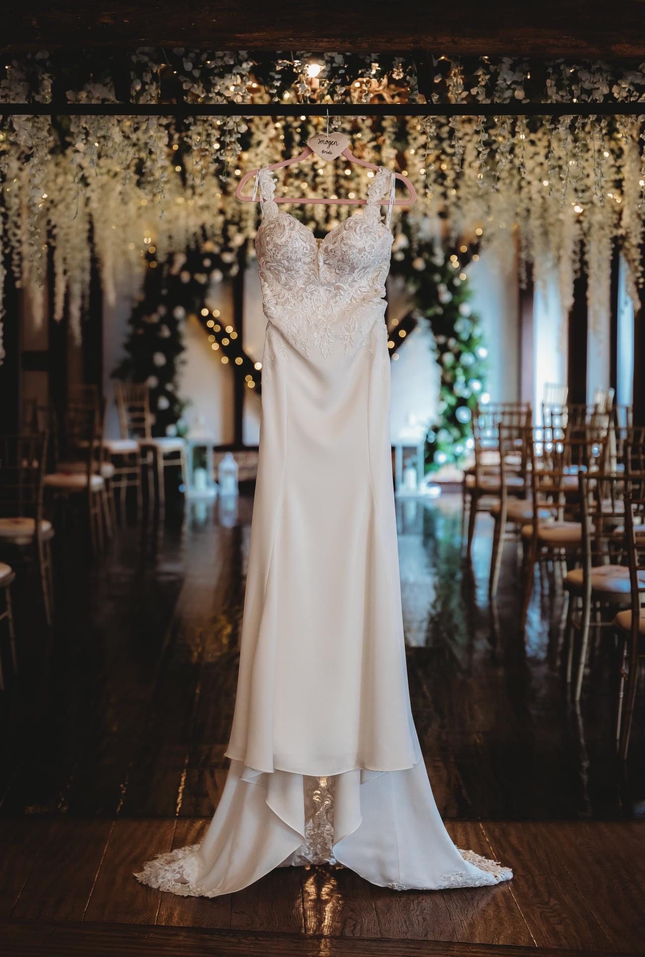 An elegant white wedding dress hangs in a venue decorated with twinkling lights and floral ceiling arrangements.
