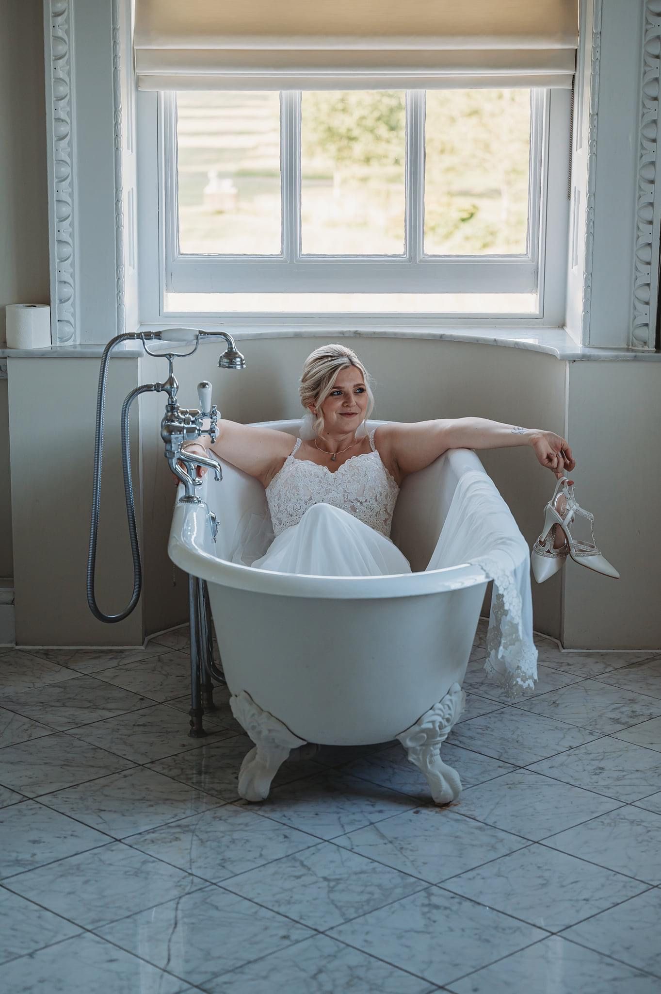 A wedding dress-clad figure relaxes in an elegant clawfoot bathtub near a bright window.
