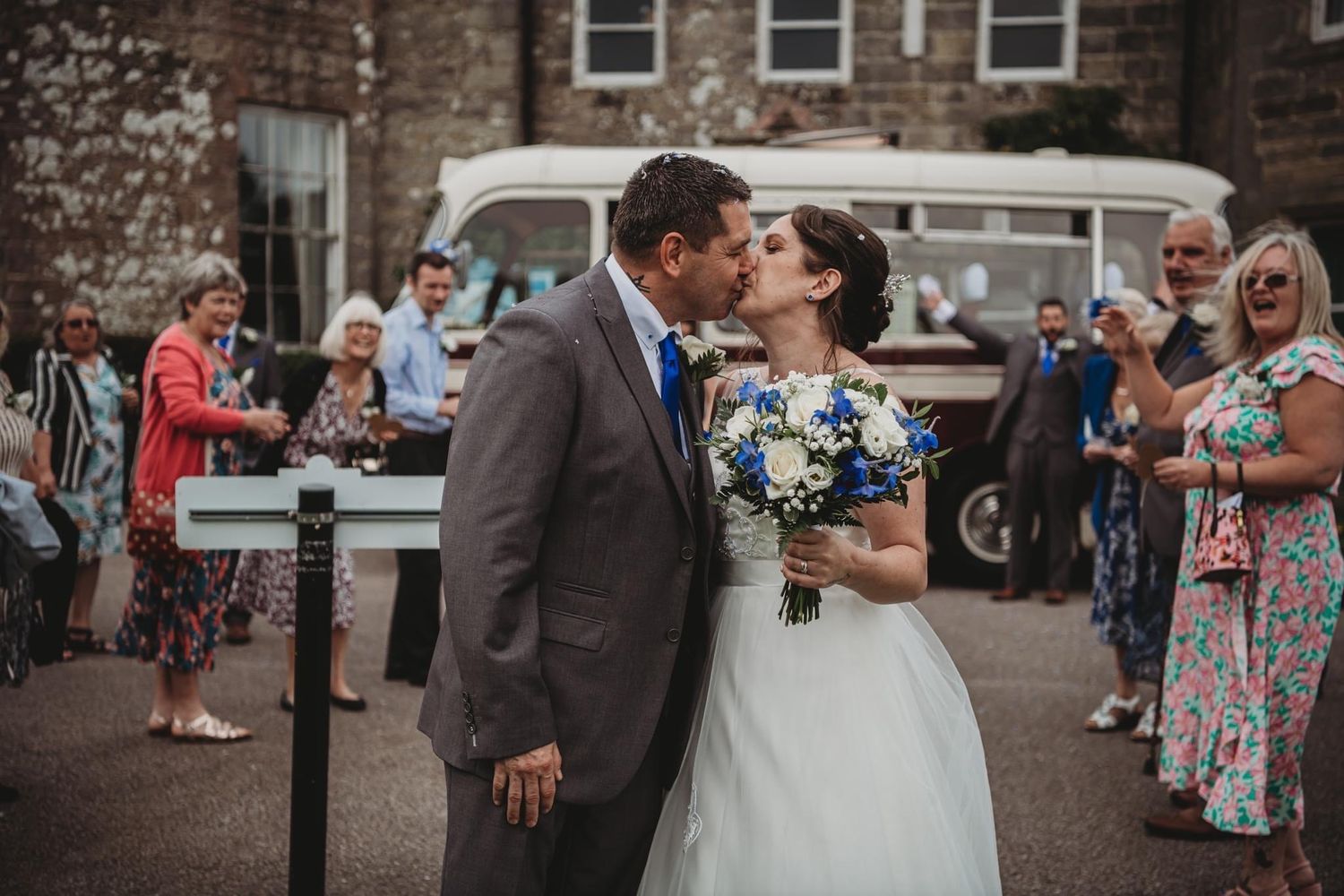 Wedding couple kisses outside a stone building while guests look on near a vintage van.