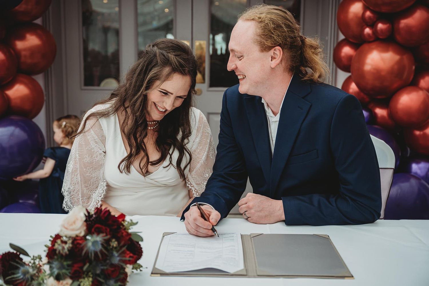 Couple signs marriage documents at a table decorated with red flowers and purple balloons.