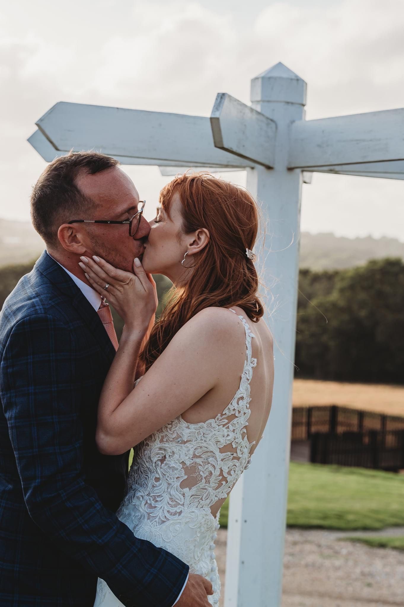 A romantic wedding kiss by a white wooden signpost in a countryside setting.