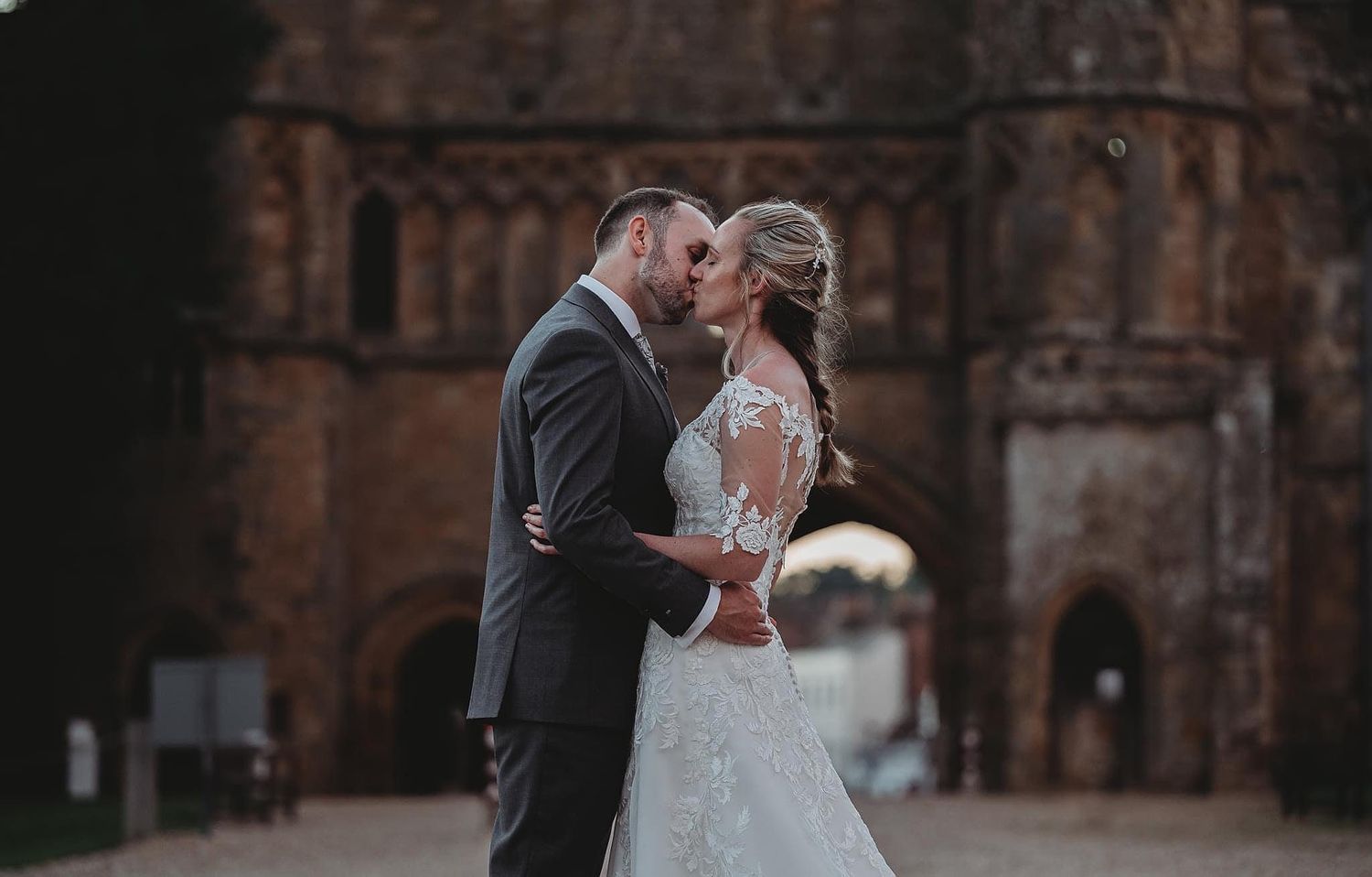 A romantic wedding moment captured in front of a historic stone building with arched architecture.
